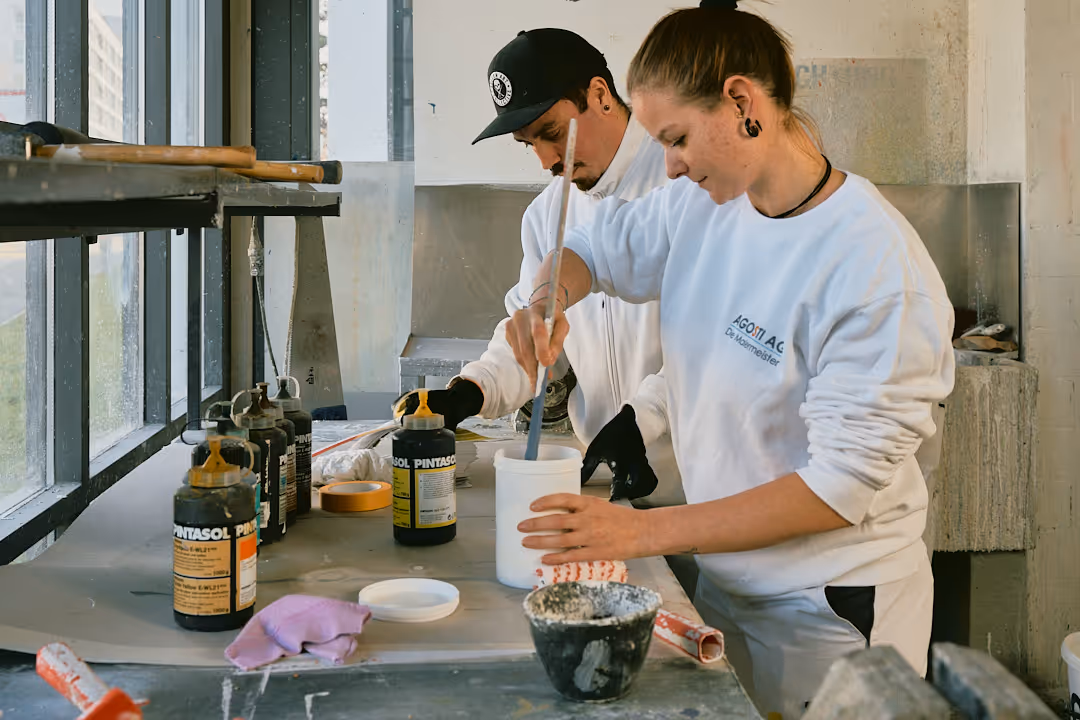 Zwei Handwerker in weisser Arbeitskleidung mischen Farbe in einem Atelier mit Flaschen und Werkzeugen auf dem Arbeitstisch.