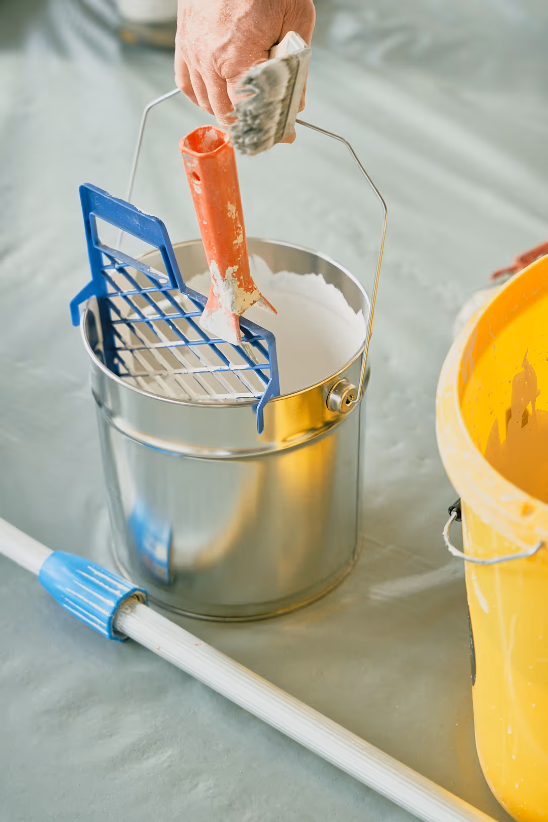 Hand hält eine Farbwanne mit weißer Farbe und einem Pinsel darüber, daneben ein gelber Eimer und eine Teleskopstange auf grauem Boden.