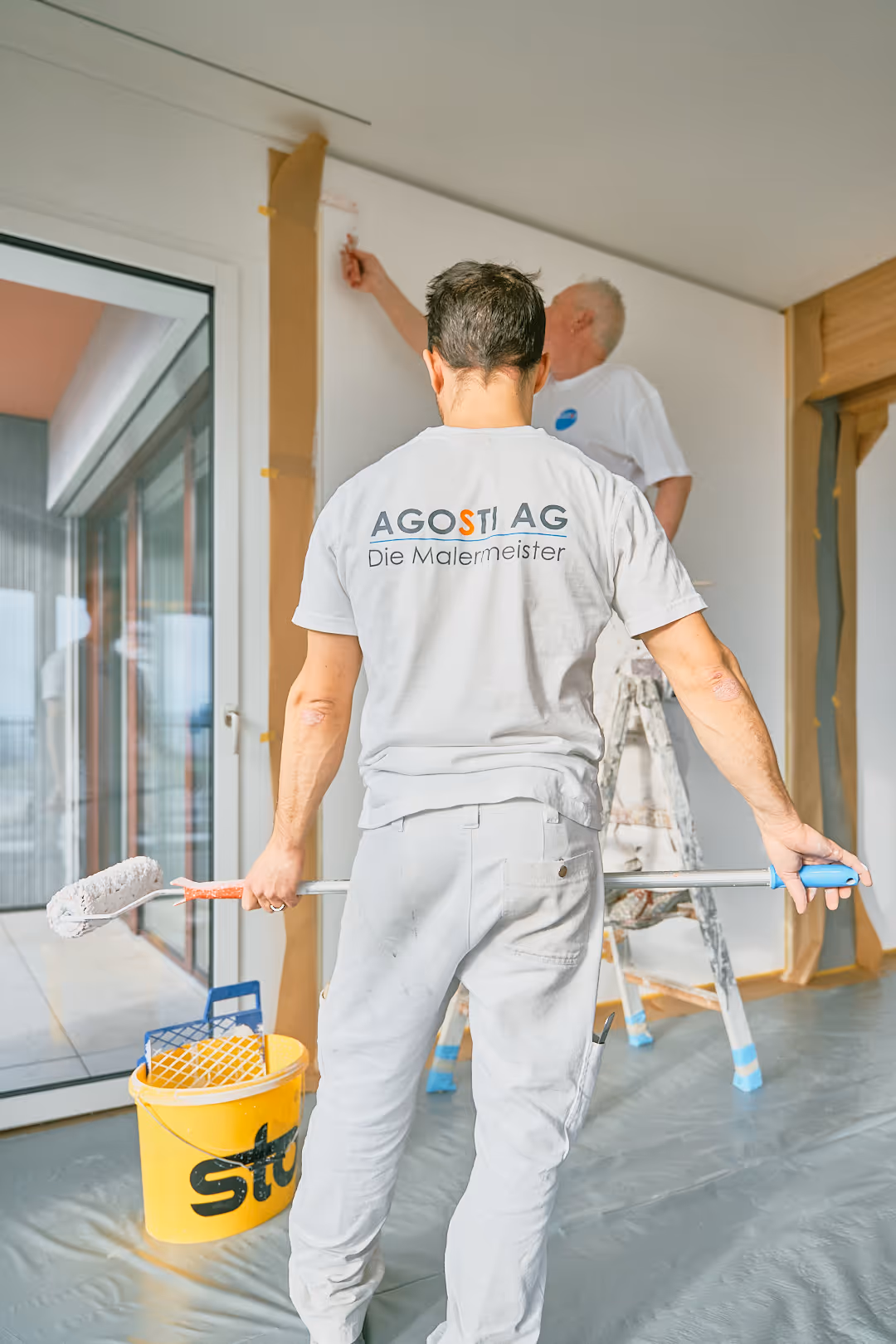 Zwei Maler in weißen Arbeitskleidern, einer mit Farbwalze vor einem Eimer, der andere malt eine Wand, in einem modern eingerichteten Raum mit großen Fenstern.