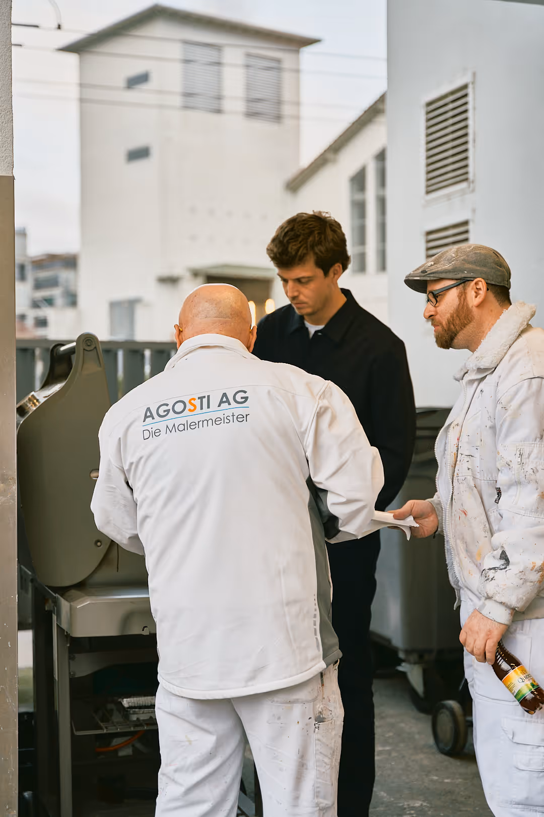 Drei Männer in Arbeitskleidung neben einem Grill, einer trägt ein Hemd mit Aufschrift AGOSTI AG Die Malermeister.