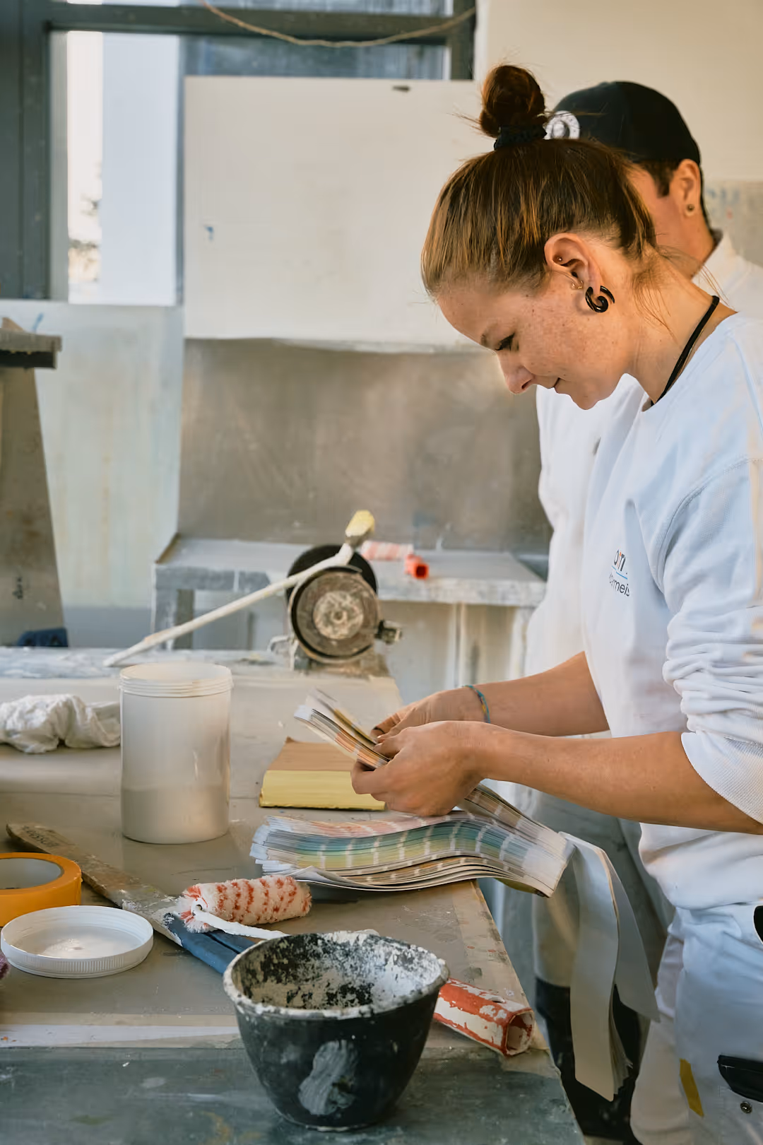 Junge Frau in Arbeitskleidung betrachtet Farbmuster in einem Werkraum mit Malerwerkzeugen auf dem Tisch.