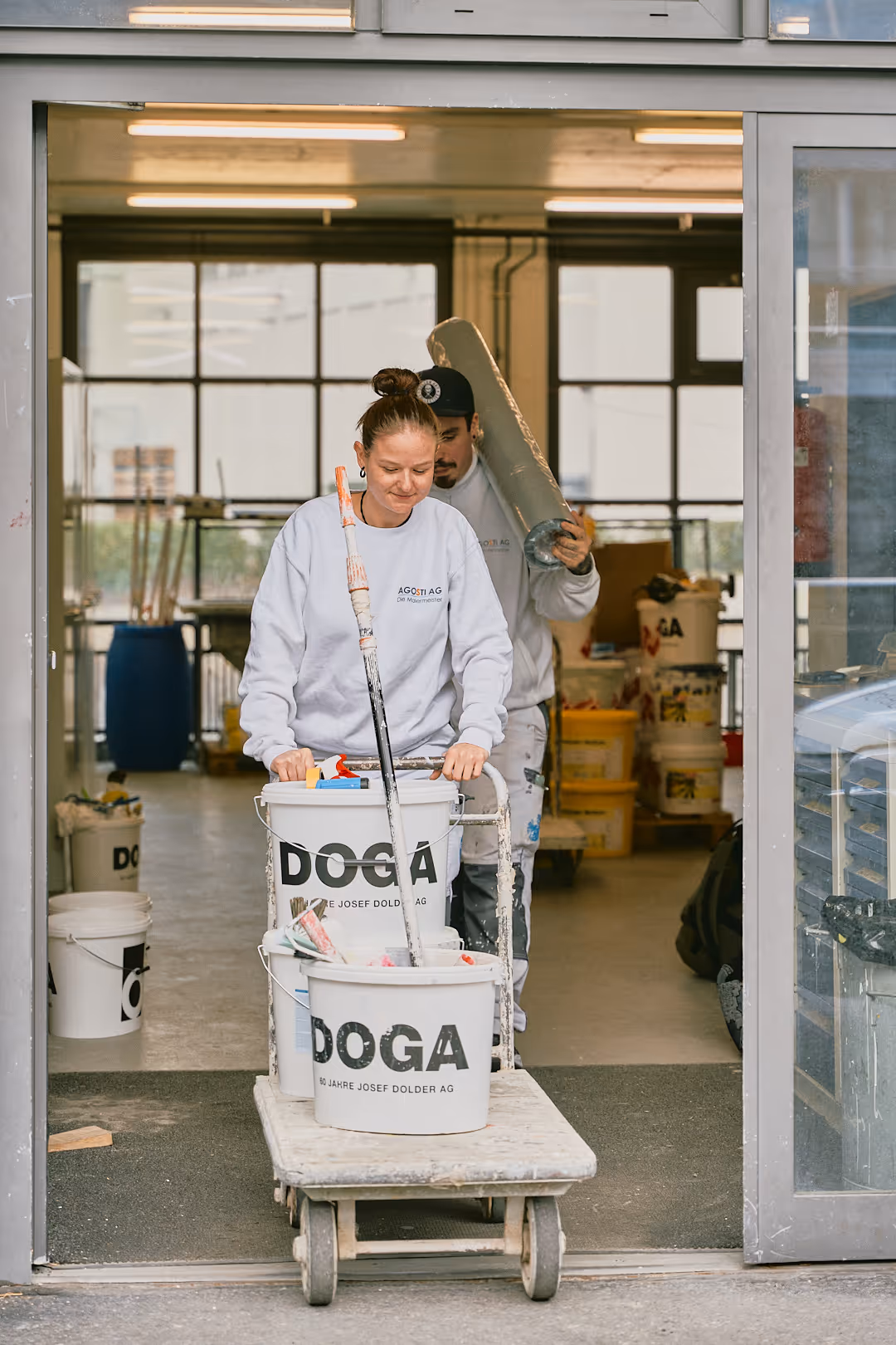 Zwei Arbeitende in Weissen Pullis, eine Frau schiebt eine Plywood-Palette mit Farbeimern, ein Mann trägt eine Rolle Isoliermaterial.