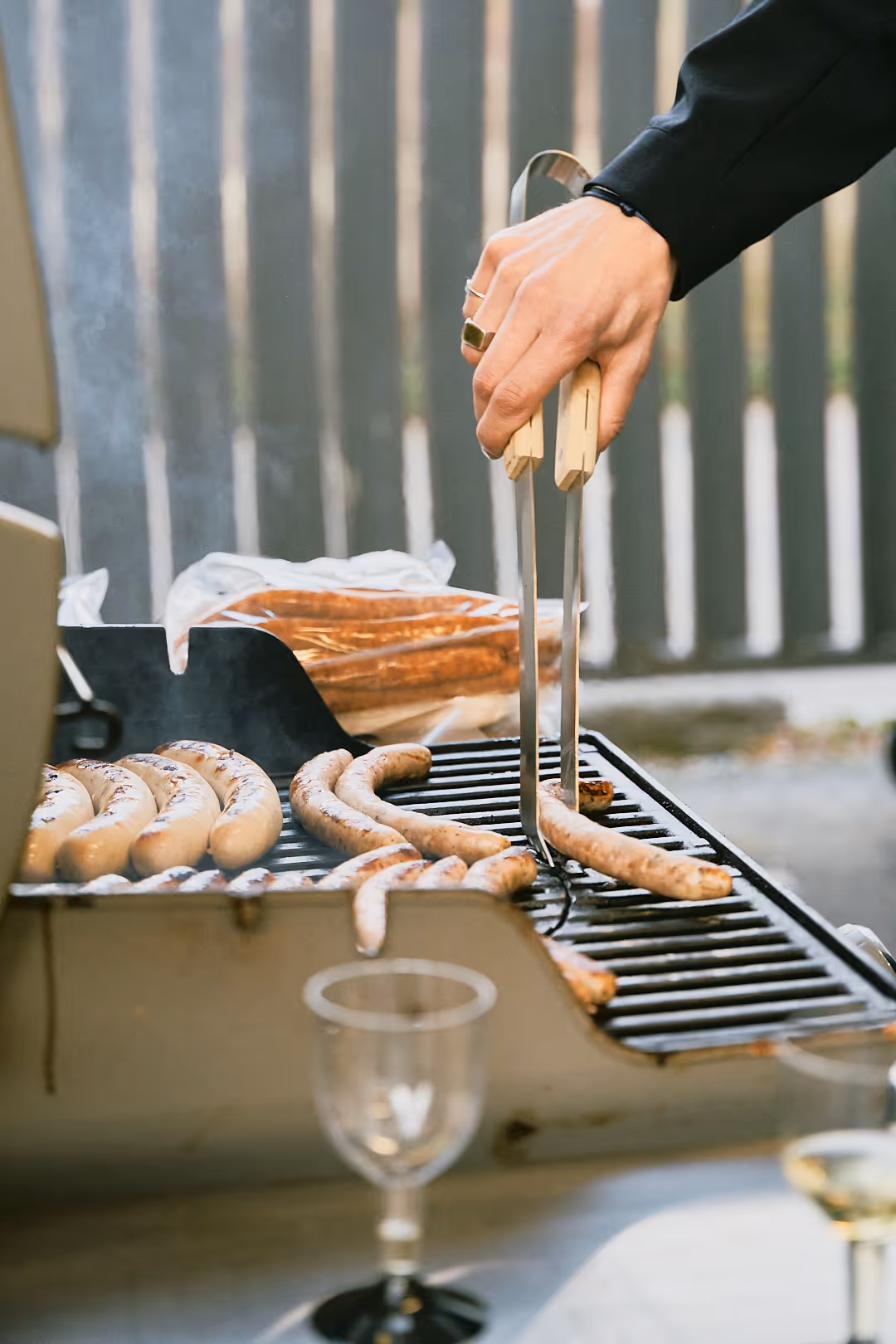 Hand mit Grillzange wendet Bratwürste auf einem Grill im Freien.