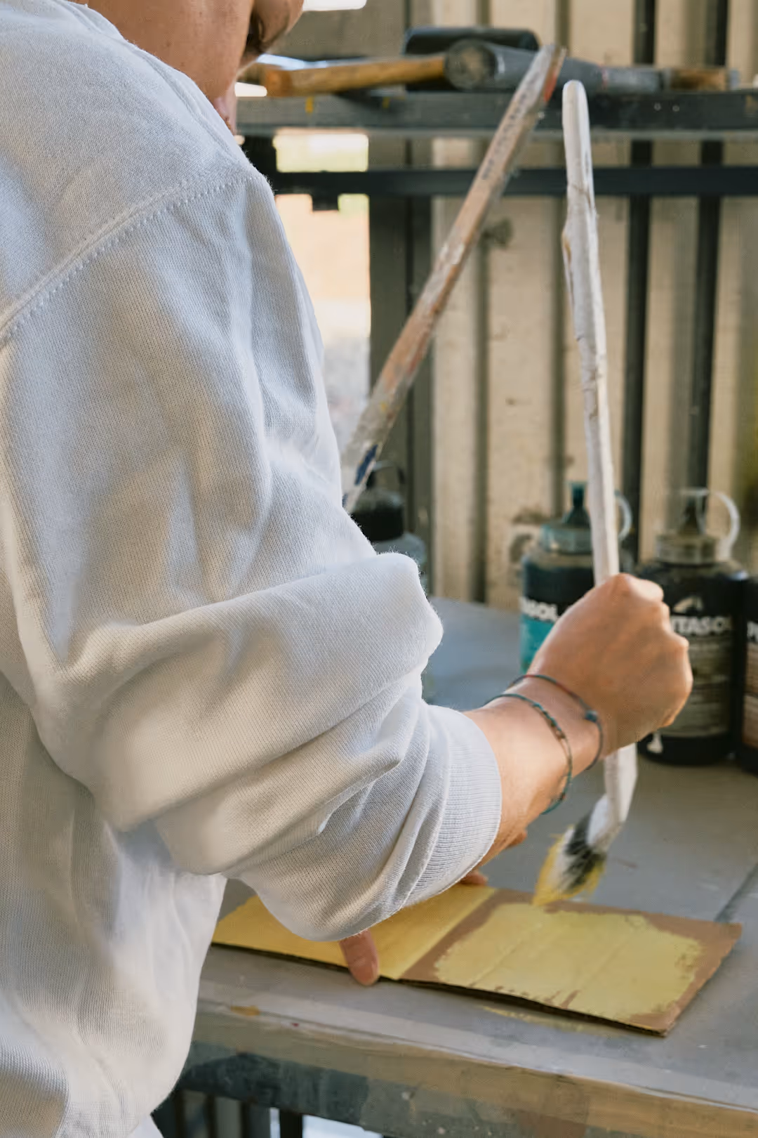Person mit weissem Pullover malt mit breitem Pinsel gelbe Farbe auf eine Holzplatte.