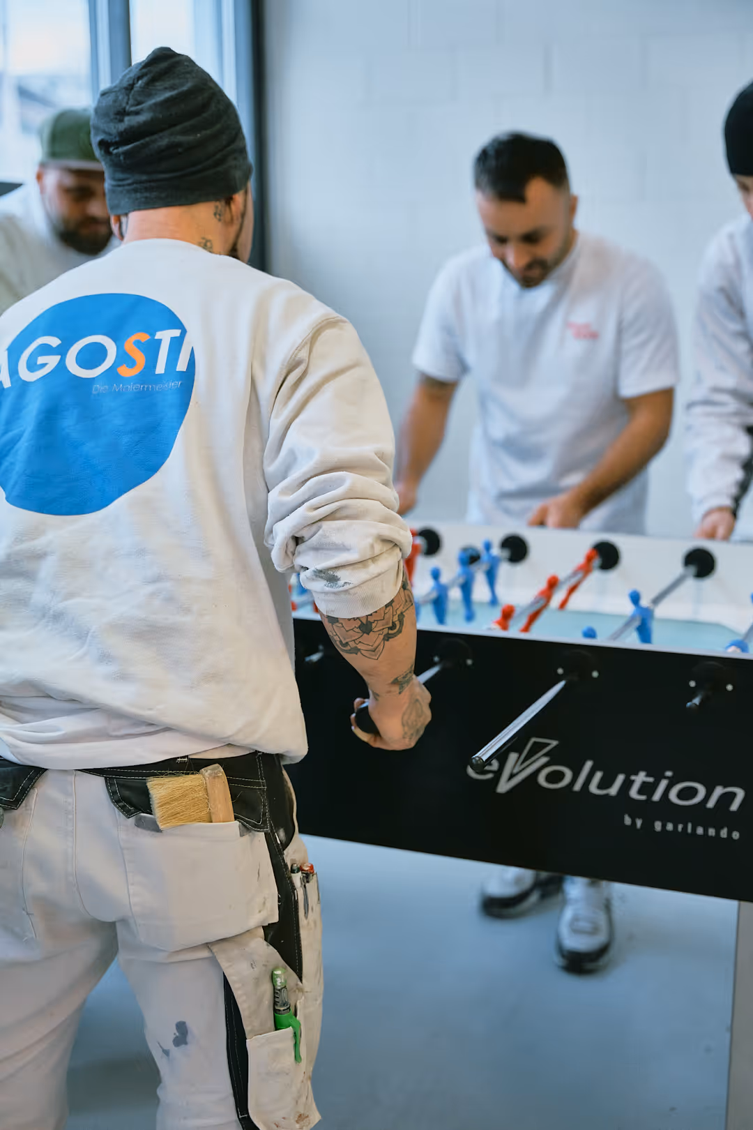 Person mit Arbeitskleidung und Werkzeugen in den Taschen spielt Tischfussball gegen zwei weitere Männer in einem hellen Raum.