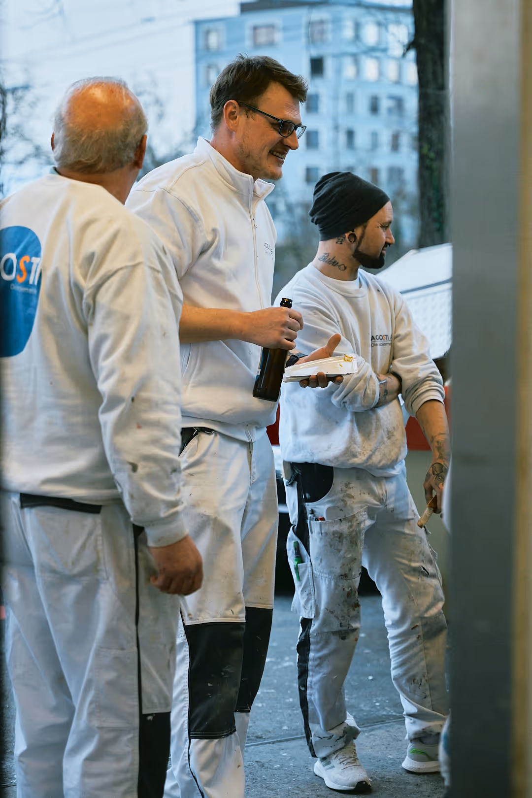 Drei Handwerker in schmutziger weißer Arbeitskleidung stehen entspannt draussen, einer hält ein Getränk und einen Teller mit Essen.