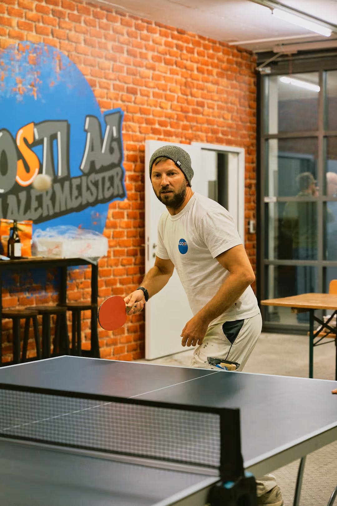 Mann mit grauer Mütze und weissem T-Shirt spielt Tischtennis vor einer Backsteinwand mit bunter Wandgrafik.
