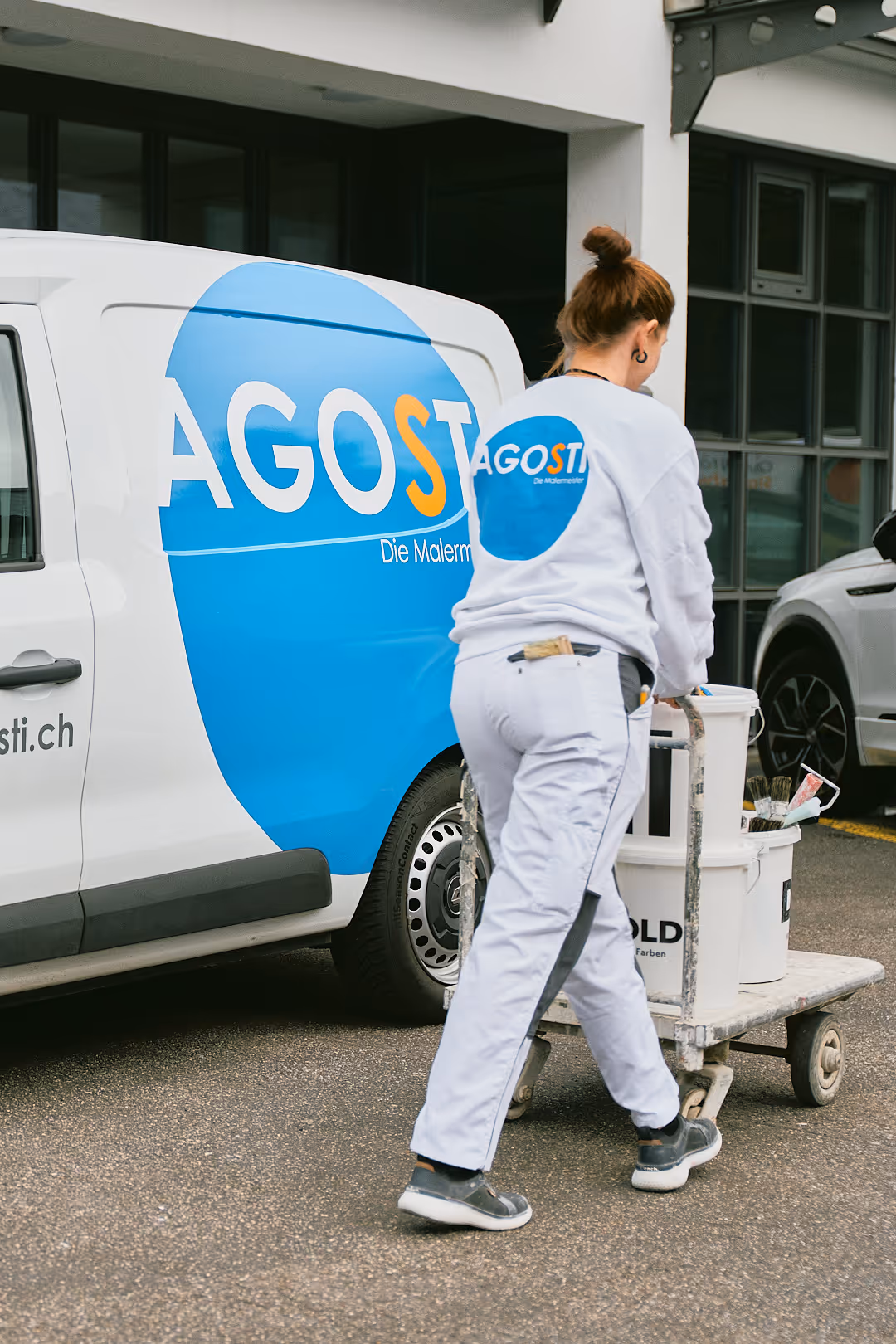 Ein Malermeister schiebt einen Wagen mit Farbeimern vor einem AGOSTI-Firmenwagen.