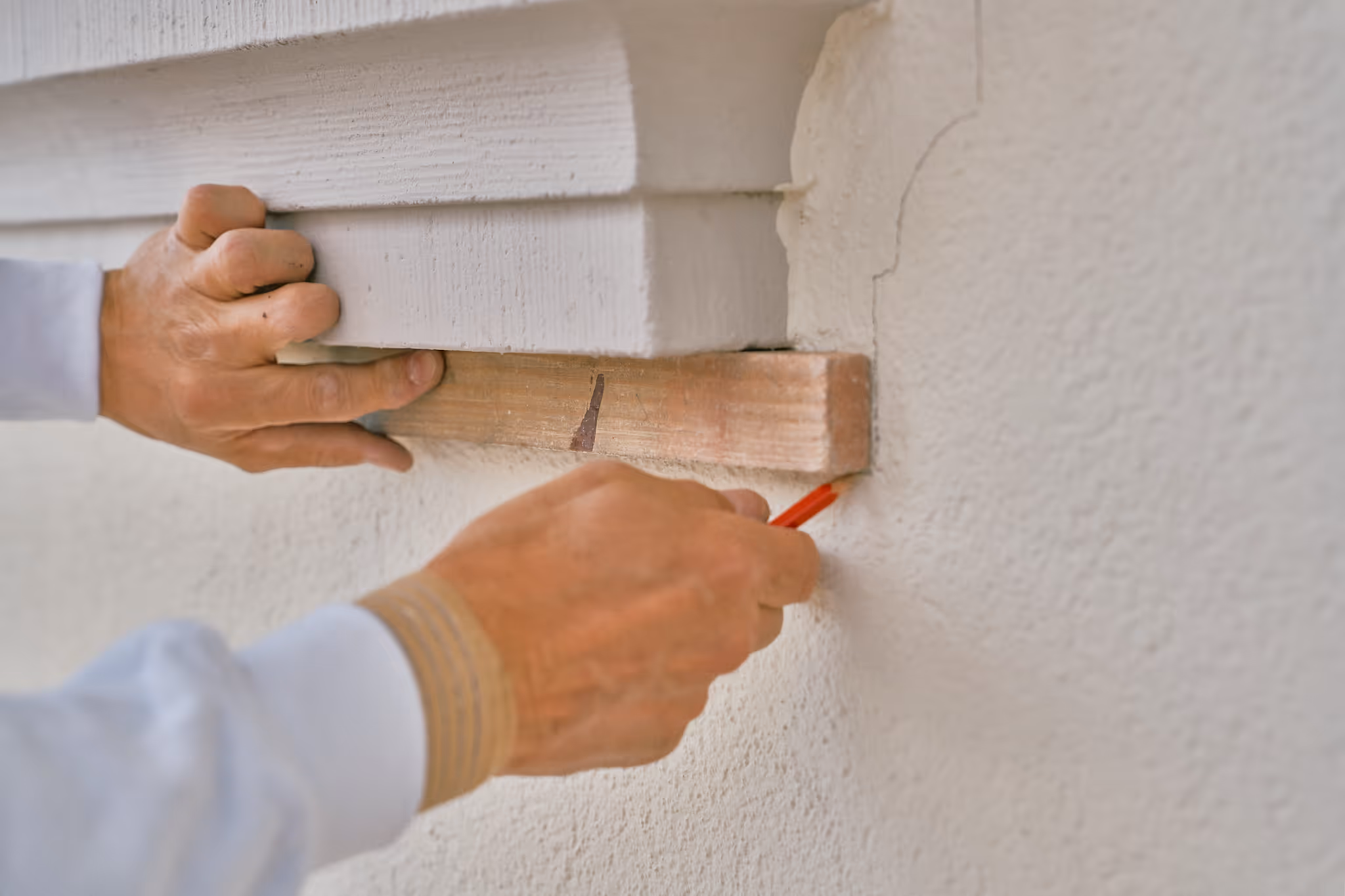 Person markiert mit einem roten Bleistift eine Linie an einer Wand mit Hilfe eines Holzlattenstücks.
