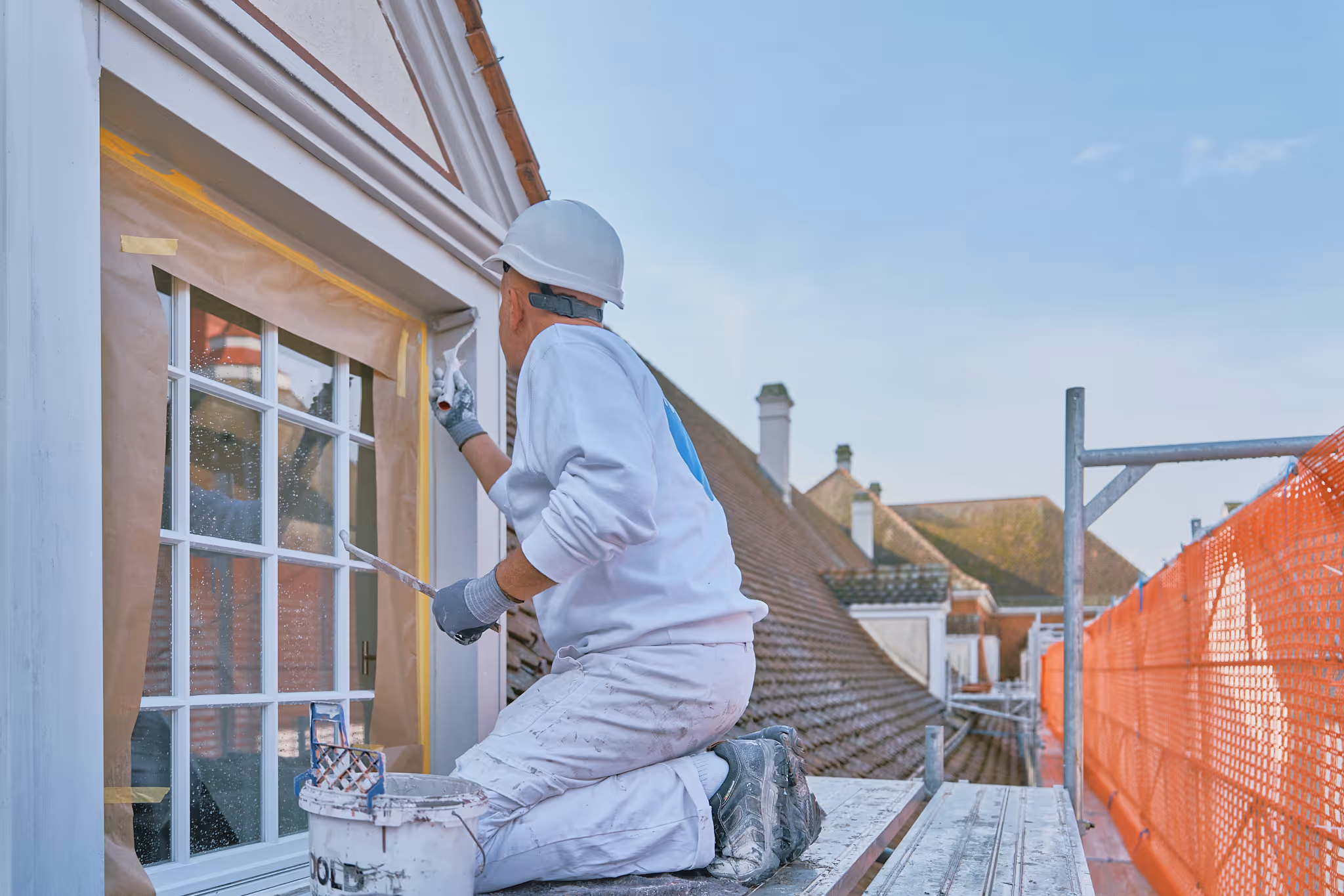 Maler in weisser Arbeitskleidung und Helm streicht ein Fenster an einem Hausdach, auf einem Gerüst mit oranger Sicherheitsabsperrung.