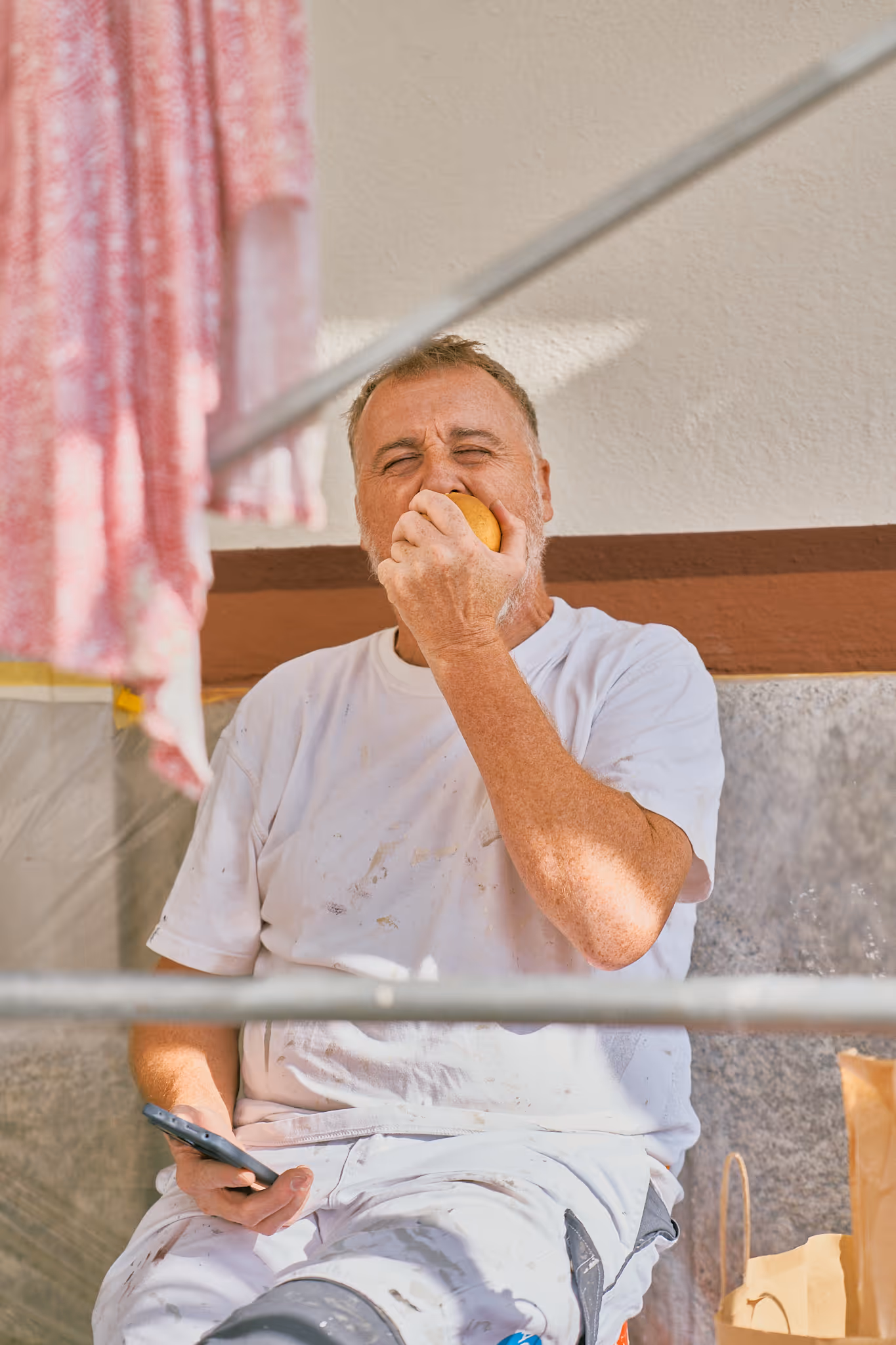 Mann in weißer Arbeitskleidung macht Pause und isst eine Orange, während er ein Smartphone hält.