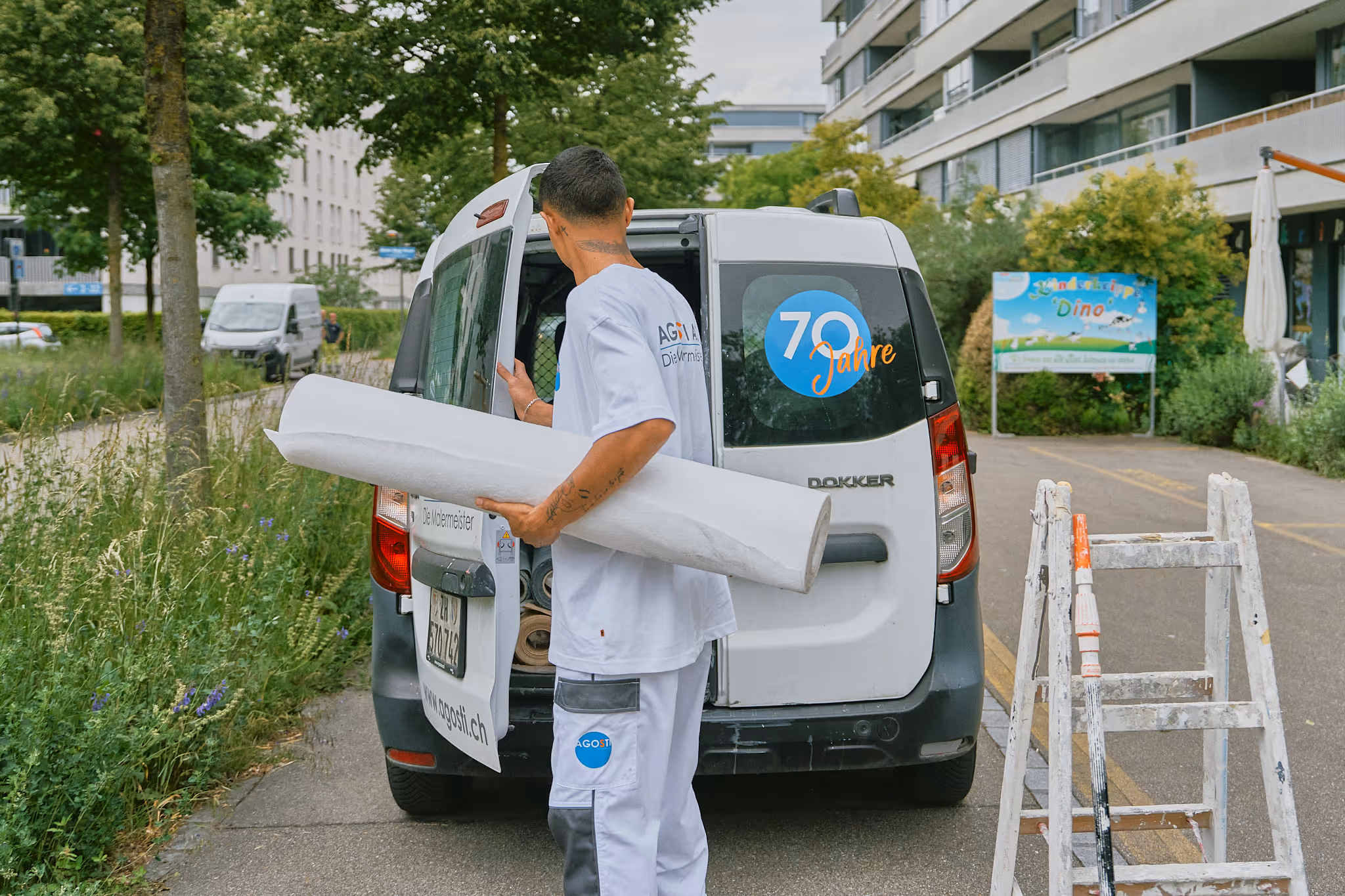 Mann in weisser Arbeitskleidung mit Rolle Papier vor einem weissen Lieferwagen mit Aufschrift '70 Jahre' auf der Heckscheibe.