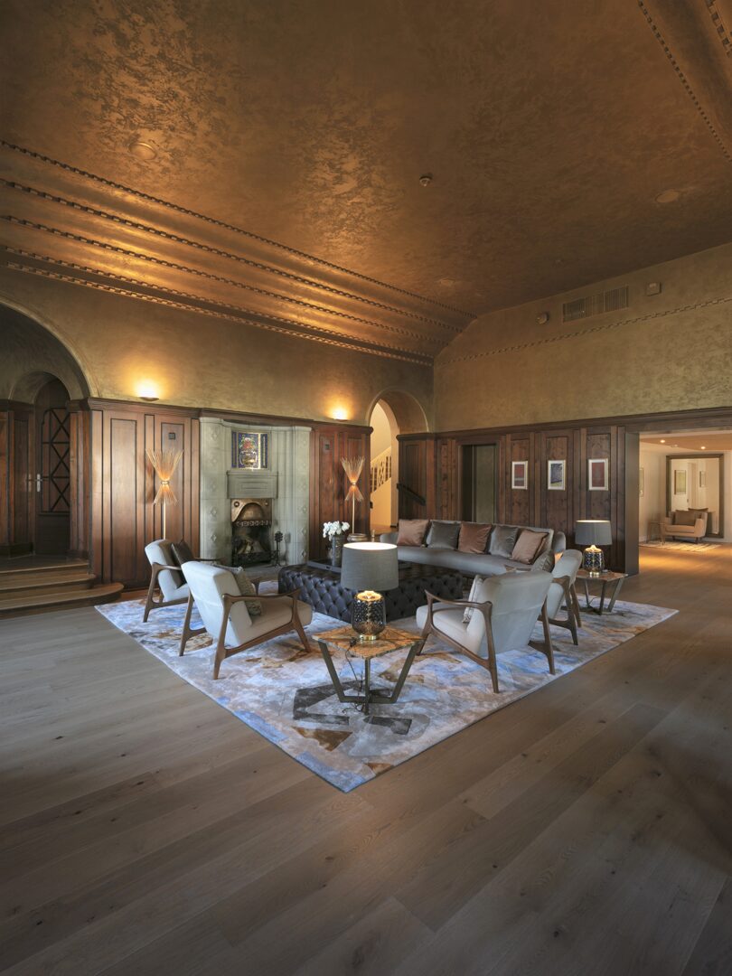 Elegantes Wohnzimmer mit brauner Holzvertäfelung, Kamin, einem großen Teppich und Sitzgruppe mit grauen Sesseln und braunem Sofa.