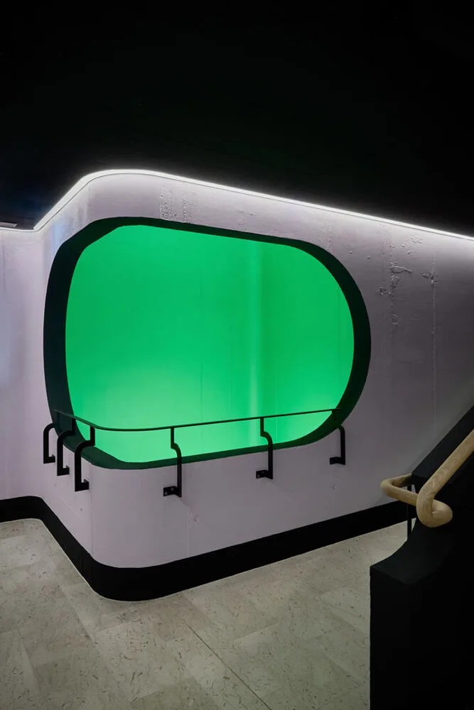 Modernes Interieur mit einer beleuchteten grünen Fensteröffnung in einer weißen Wand mit schwarzen Umrandungen und Handlauf.
