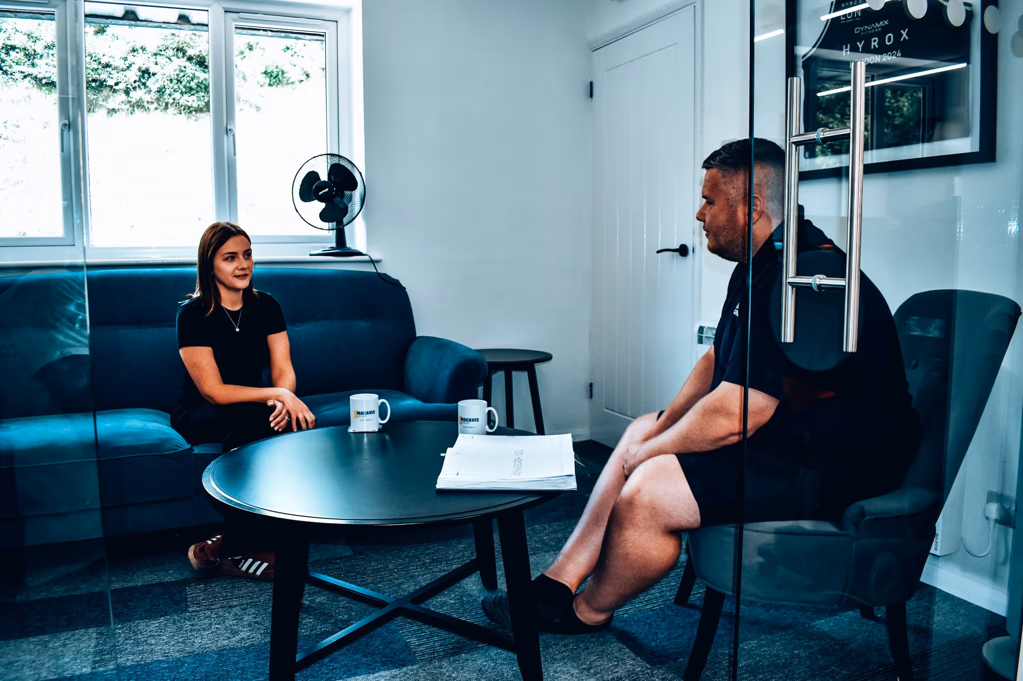 Two people in an office meeting with coffee mugs and a fan