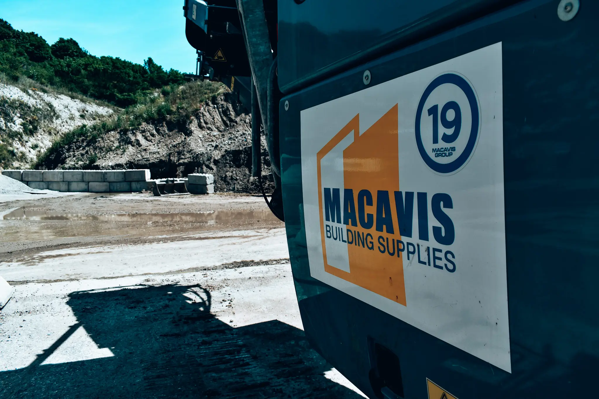 Close-up of a construction vehicle with a sign reading 'MACAVIS Building Supplies' at a muddy construction site with concrete blocks and green shrub-covered hills in the background.