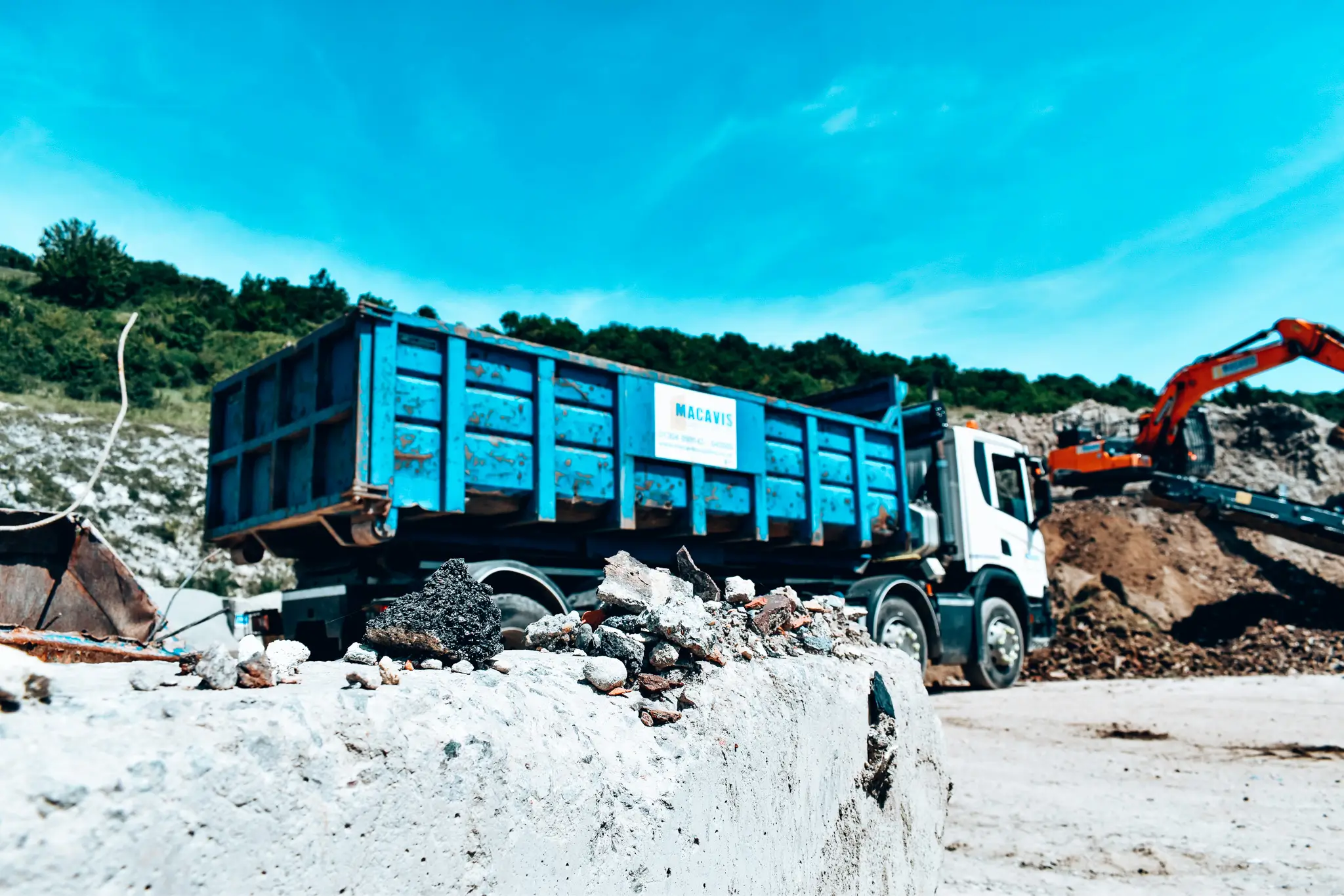Blue dump truck at a construction site with an orange excavator digging dirt in the background.