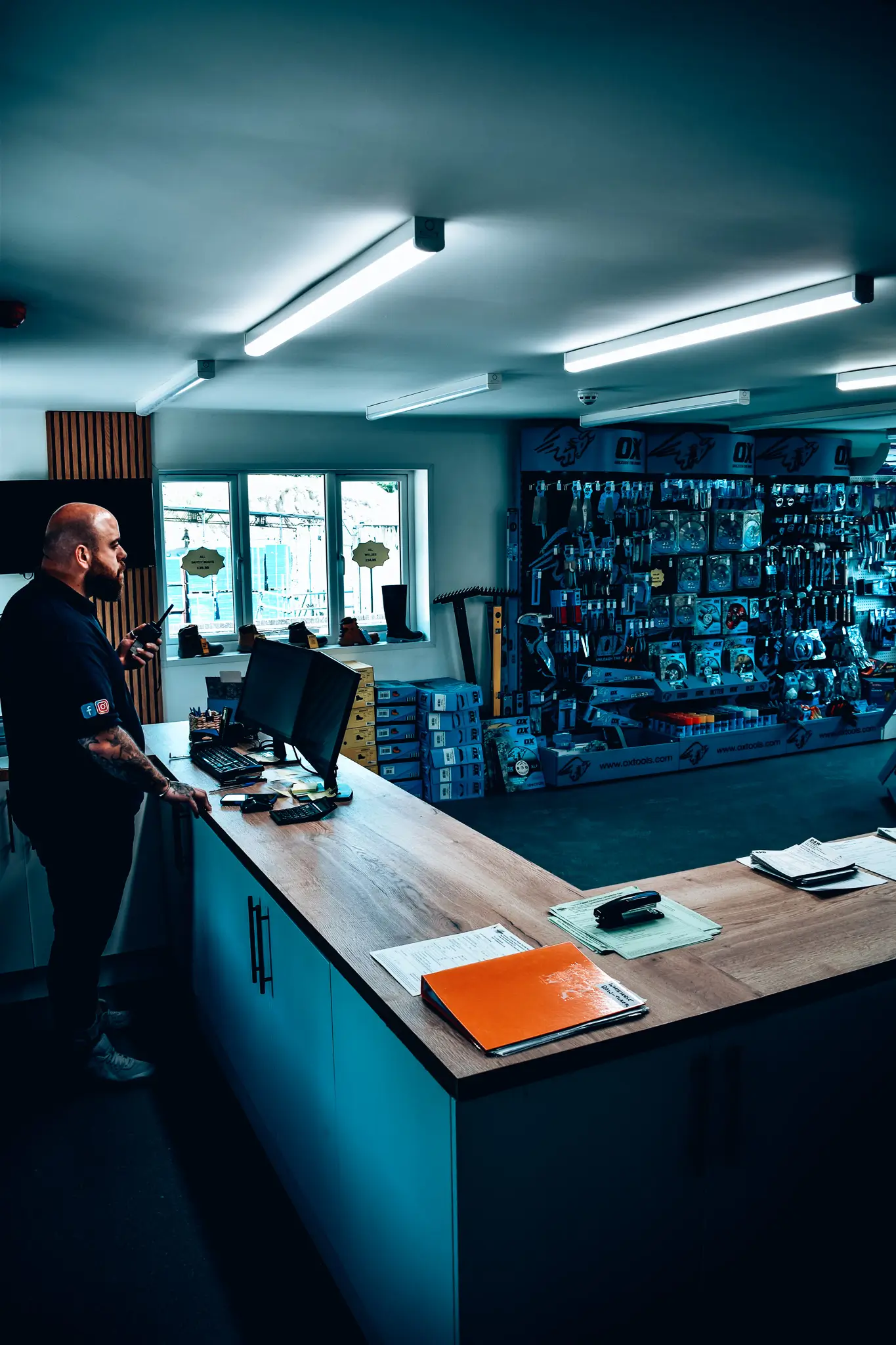 A man with tattoos stands behind a wooden counter in a hardware store, speaking into a walkie-talkie with various tools and equipment displayed on shelves behind him.