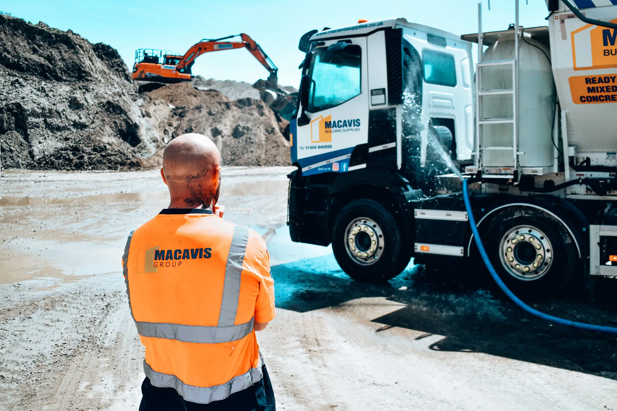 Man with tattoos wearing a high-visibility Macavis Group vest stands near a Macavis cement mixer truck at a muddy construction site with an excavator in the background.