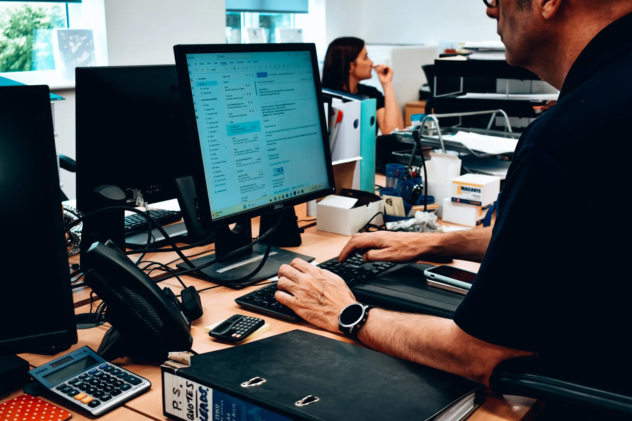 Man typing on a keyboard in an office with a computer monitor showing emails and a woman sitting in the background.