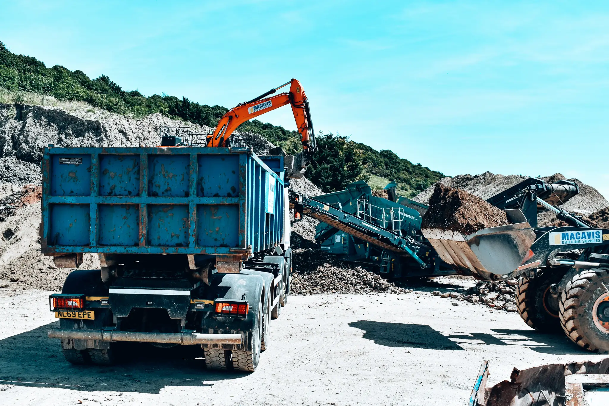 Construction site with an orange excavator loading dirt into a blue dump truck and other heavy machinery working nearby.