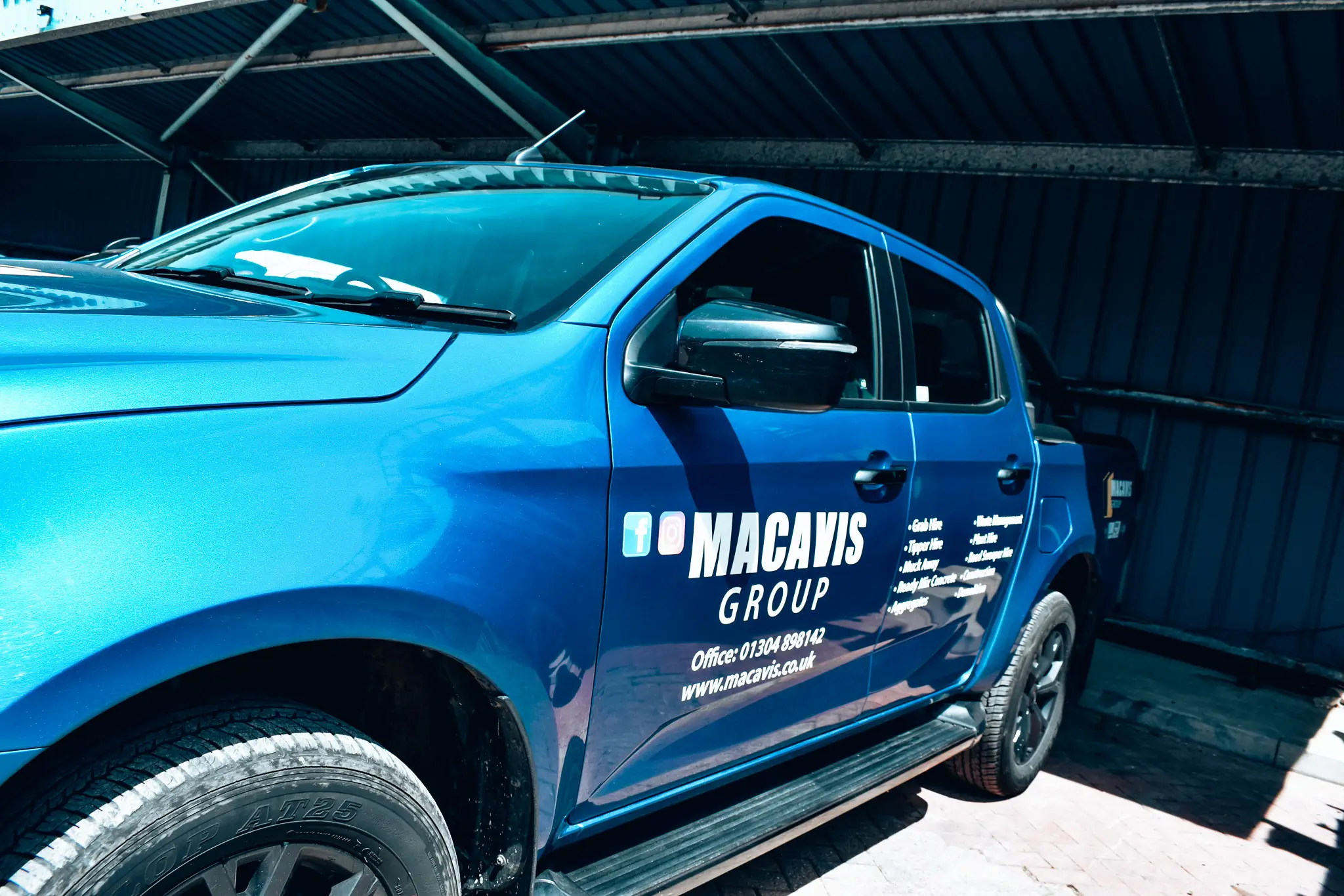 Blue pickup truck with Macavis Group company logo and contact information on the door, parked under a metal roof.