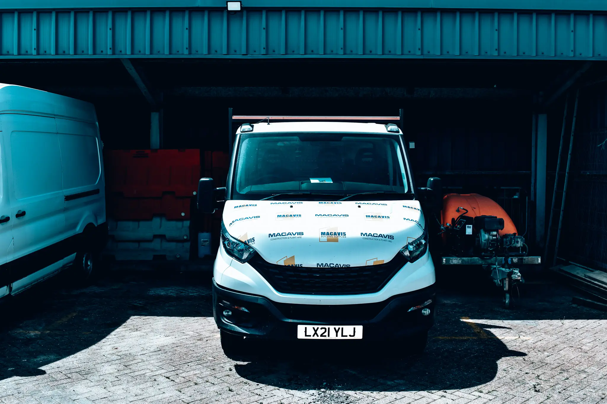 Front view of a white MacAvis-branded van with license plate LX21 YLJ parked in a shaded garage with a white van on the left and an orange boat on a trailer on the right.