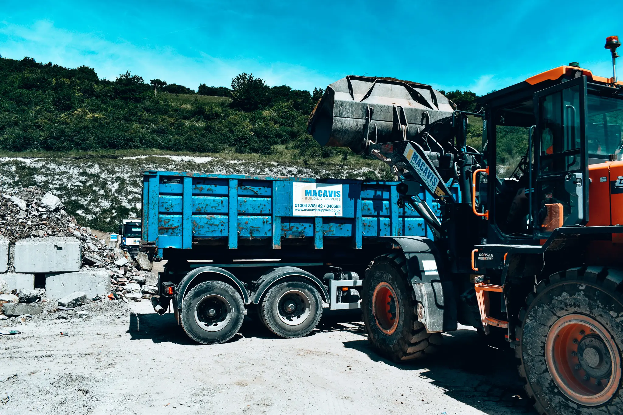 Blue MacAvis truck and orange loader at rocky construction site