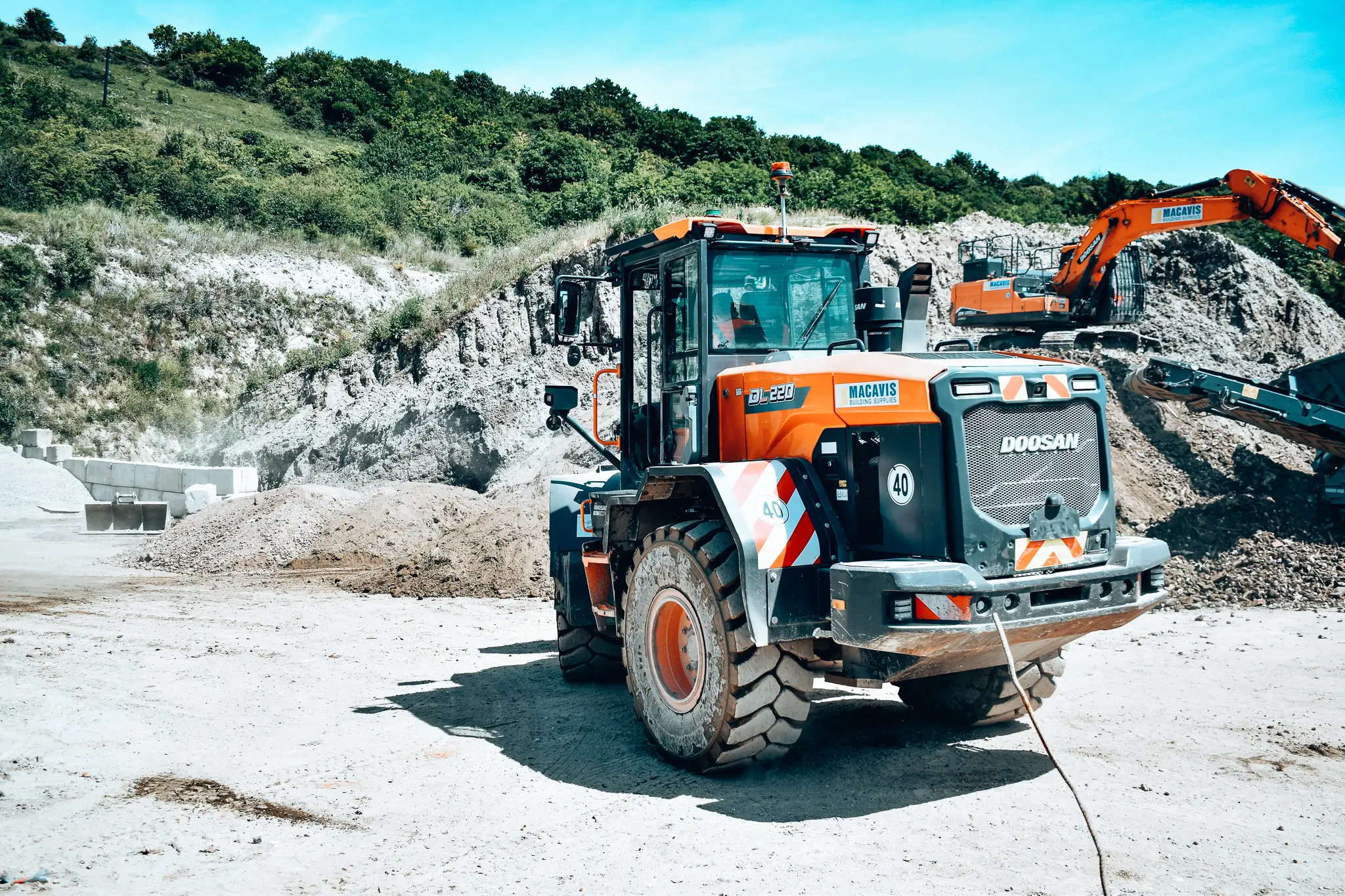 Orange Doosan DL220 construction loader parked at a dirt quarry site with excavator and conveyor in the background.