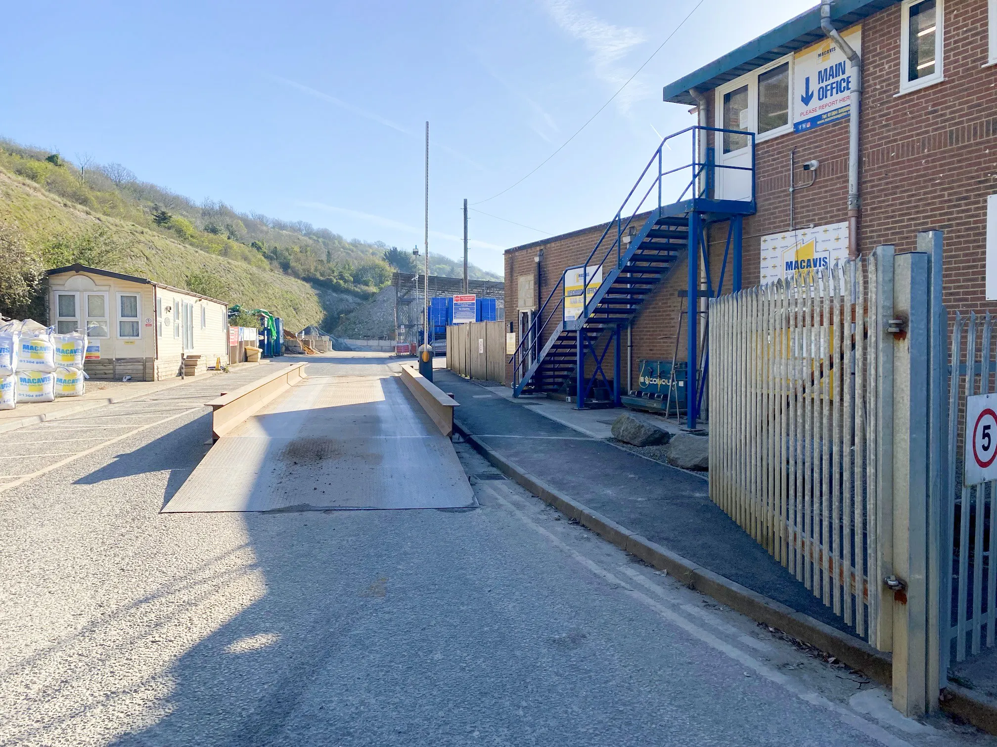Industrial entrance with metal weighbridge, blue stairs, signs, and fenced area under clear sky.