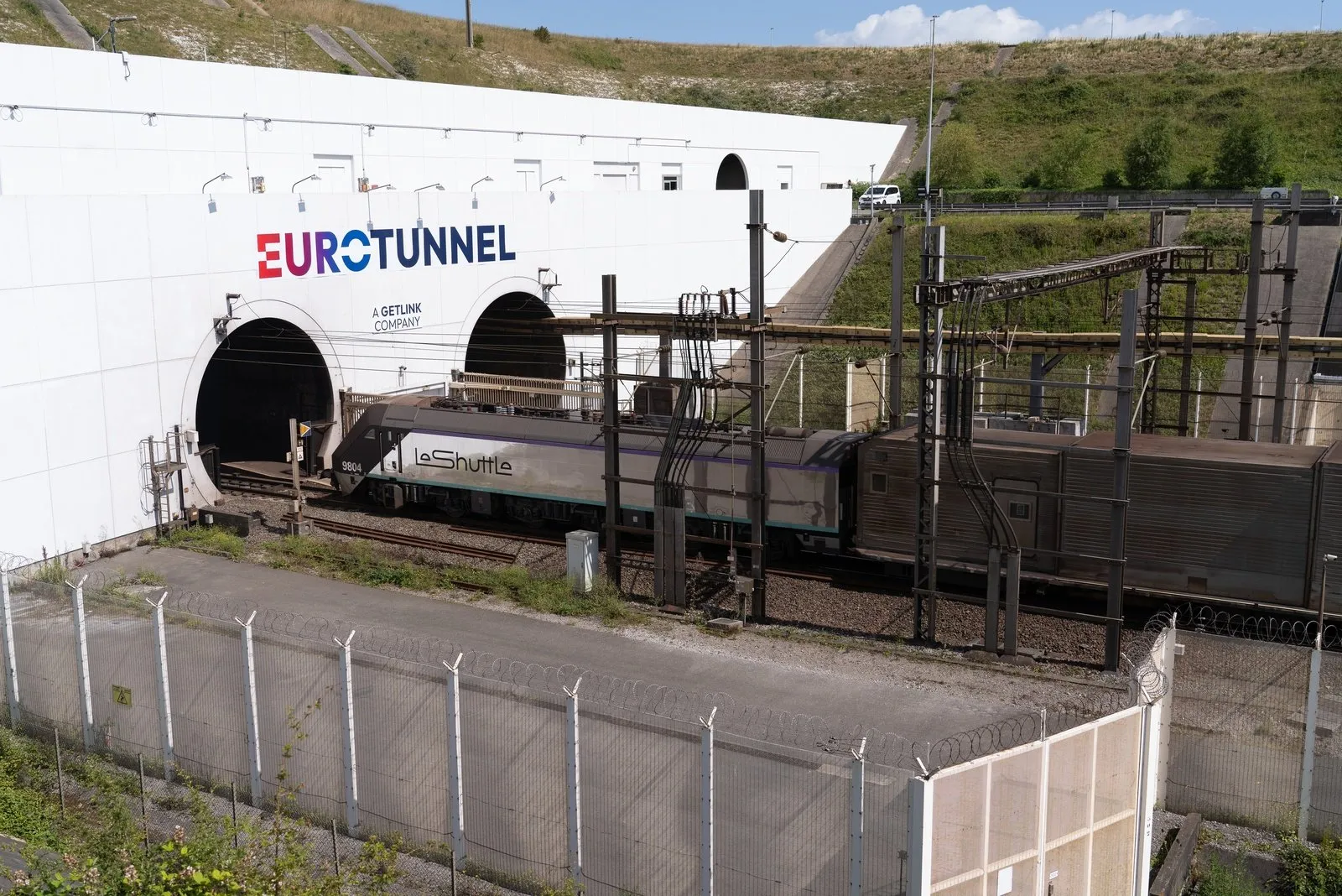 Le Shuttle train entering the Eurotunnel terminal with fences and grassy hills in the background.