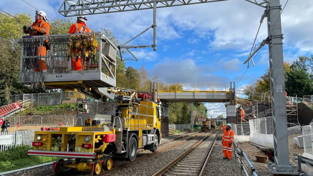 Two rail engineers in safety gear on a yellow rail vehicle working on overhead rail lines under a bridge.
