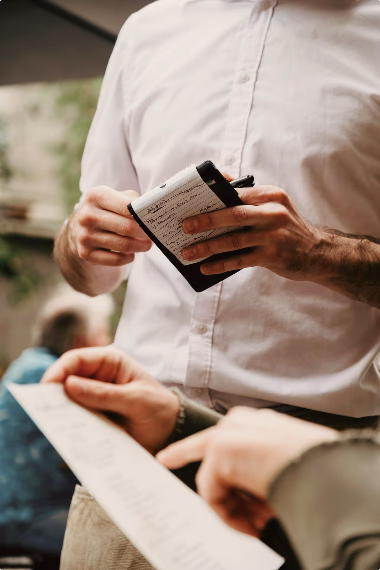 A waiter in a white shirt holds a notepad and pen, taking an order from a person holding a menu. The scene suggests a relaxed café environment.