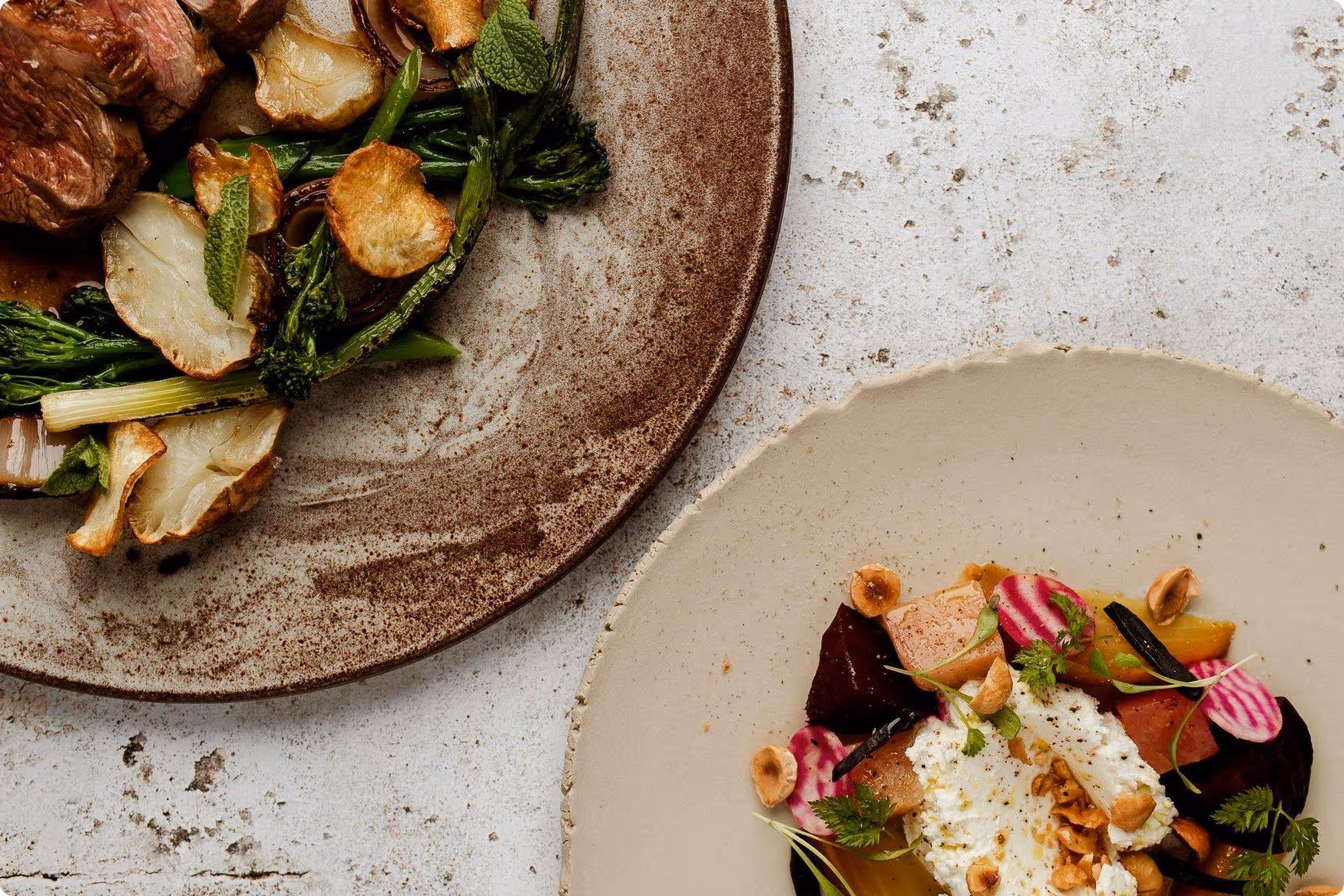 Delicate plating of a meat dish with greens and roasted roots on a rustic plate, alongside a colourful salad with beets, goat cheese, and nuts.