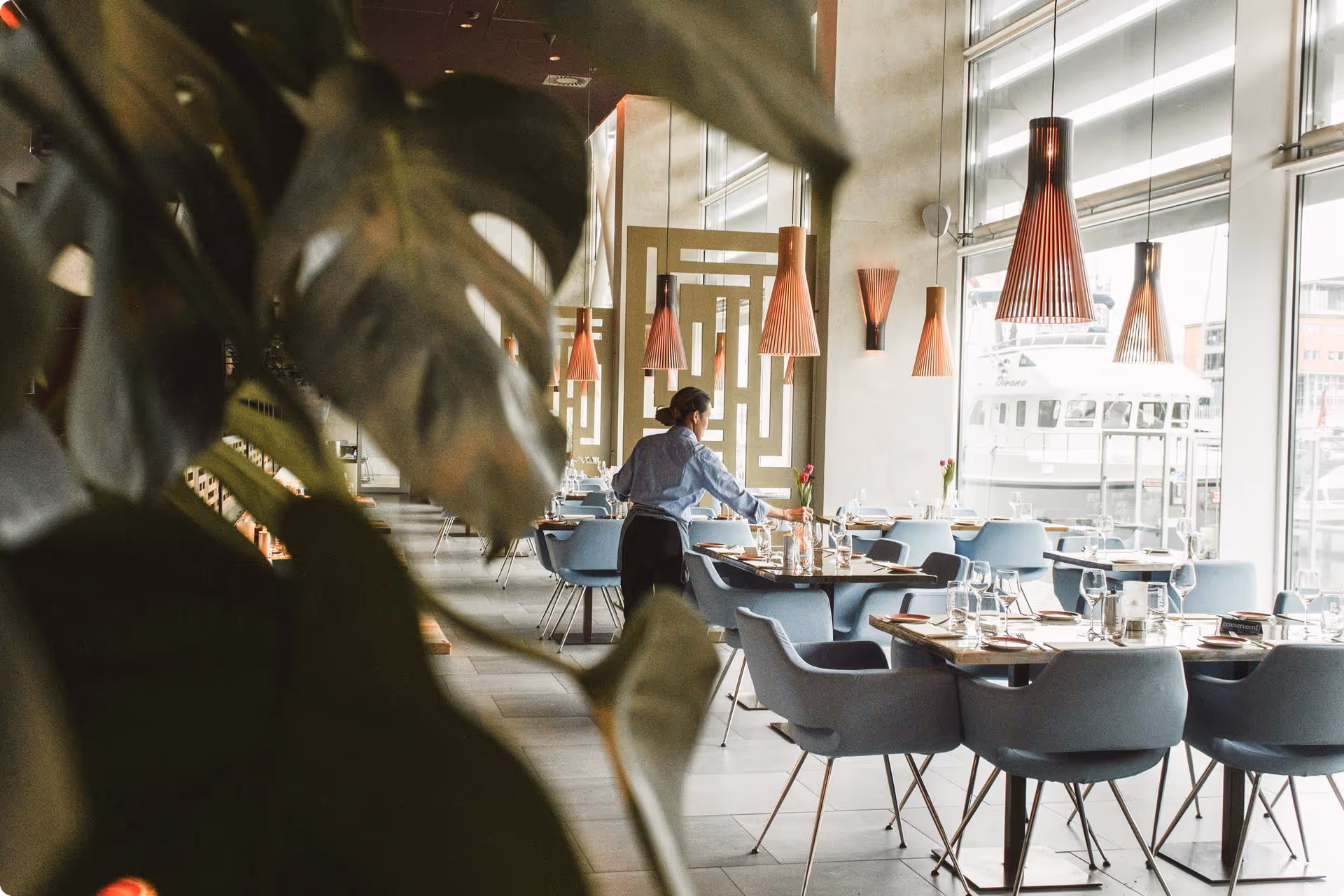 Modern restaurant interior with a calm ambiance, featuring blue chairs, set tables, and large windows. A person arranges tableware, adding to the serene atmosphere.