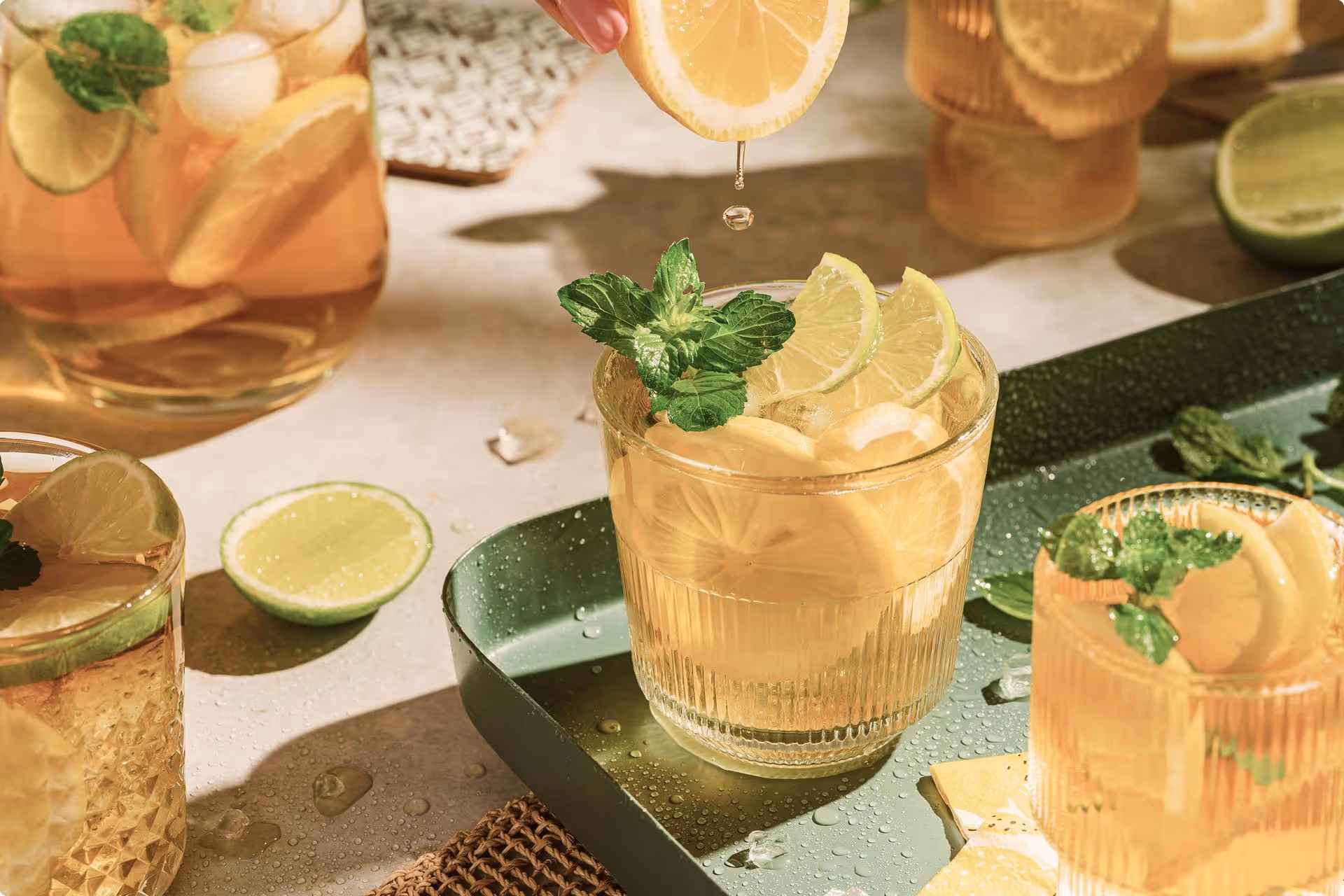Glasses of iced tea with lemon and mint on a green tray. A hand squeezes a lemon slice, adding freshness. Sunlight casts warm, summery shadows.