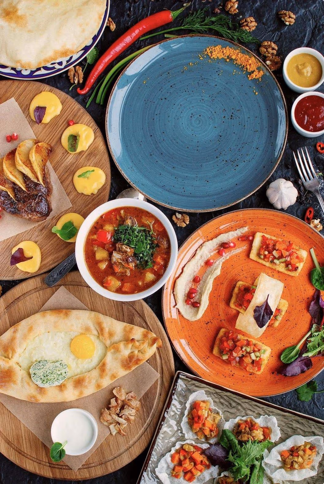 A vibrant spread of diverse dishes on colourful plates, including soup, pastry, and appetizers garnished with herbs. The scene conveys a sense of abundant, multicultural cuisine.