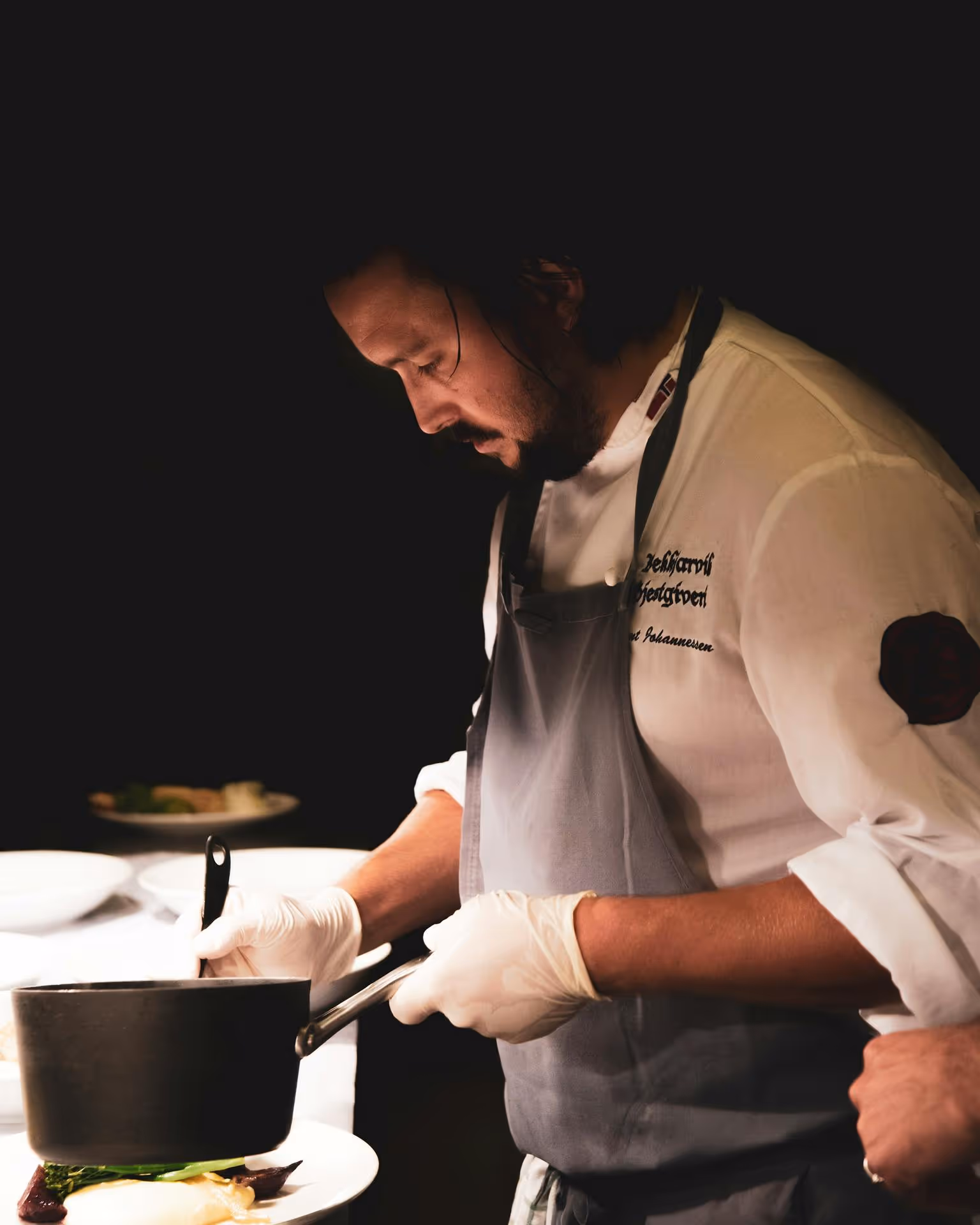 Chef in a white uniform and apron focuses intently while plating a dish under soft lighting, creating a calm, professional kitchen atmosphere.