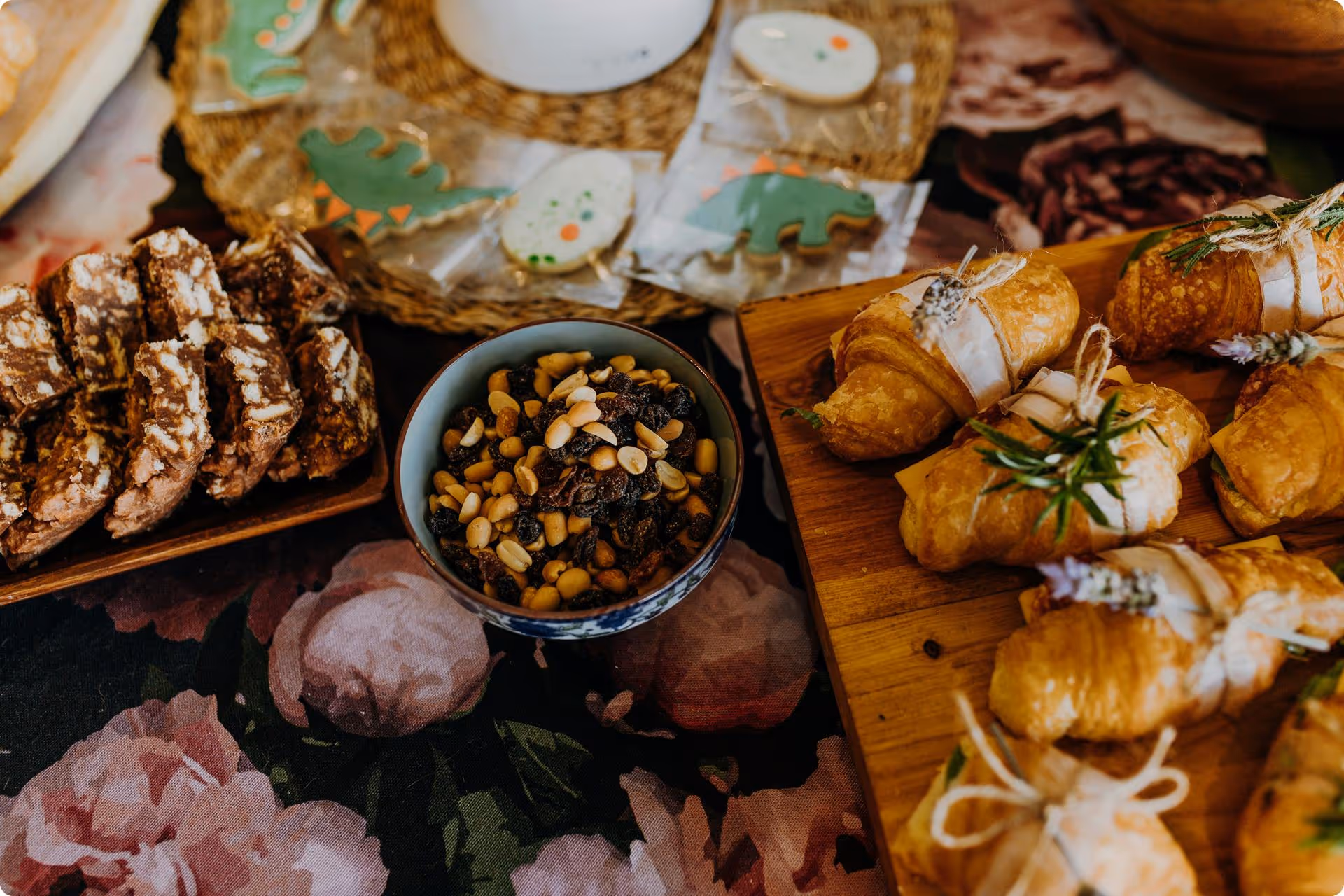 Assorted pastries and cookies on a floral tablecloth. Croissants with herbs are tied with twine, alongside a bowl of mixed nuts and dried fruit.