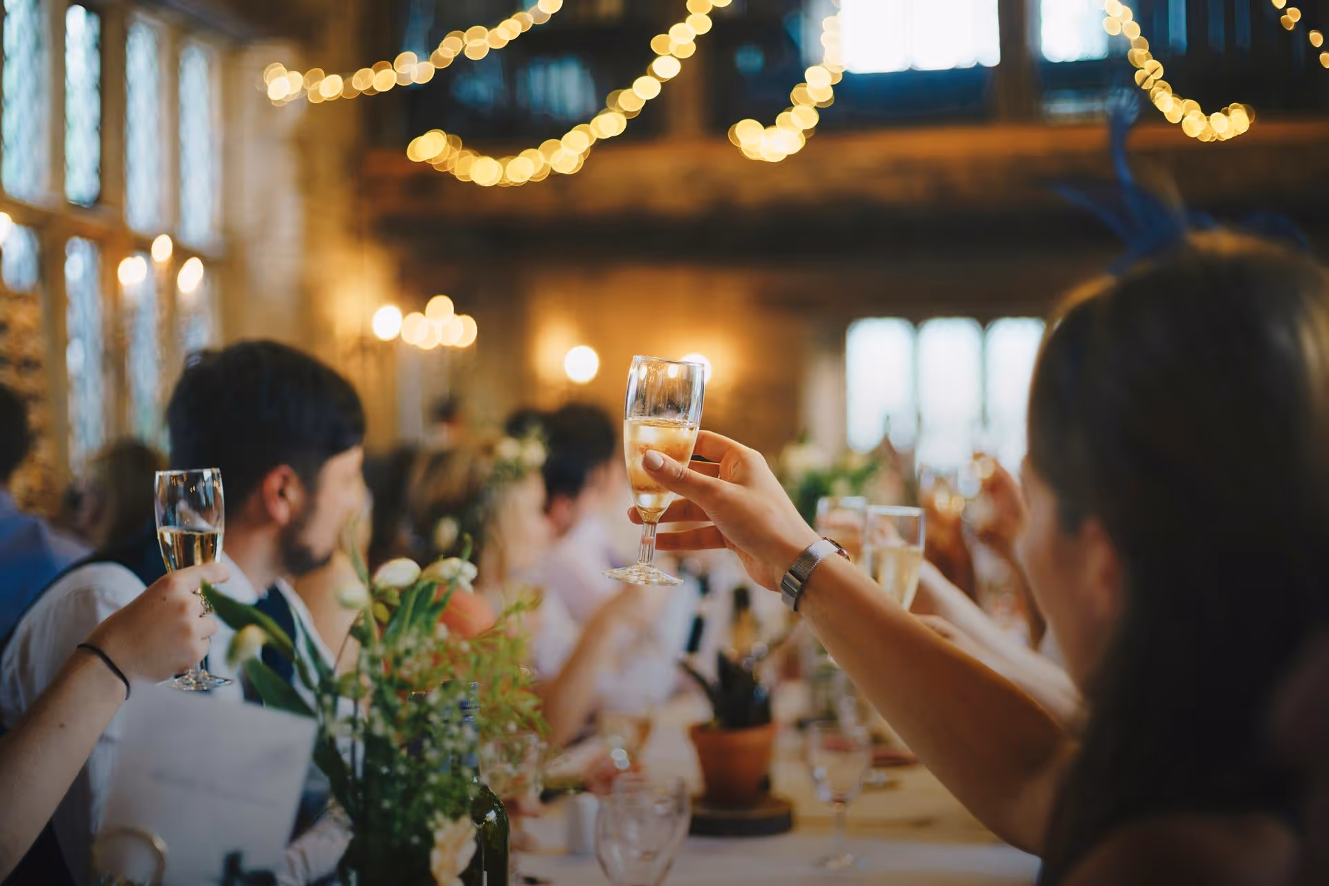 A lively celebration scene with people raising champagne glasses in a rustic venue. Warm lighting and string lights create a festive, joyful atmosphere.