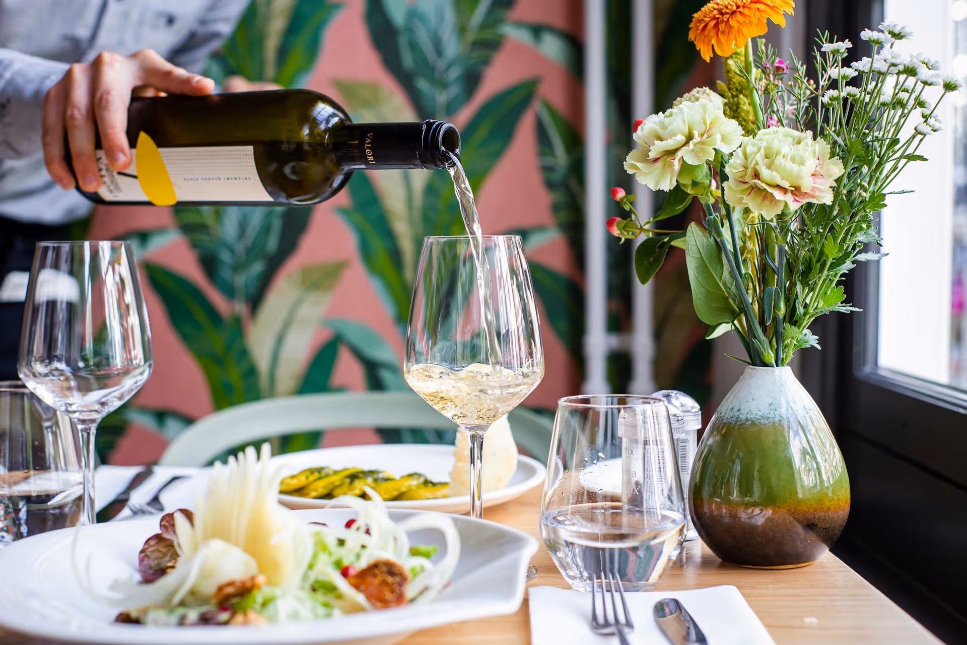 A person pours white wine into a glass on a table with elegant dishes. A vase with colorful flowers decorates the table, set against a leafy patterned wall.