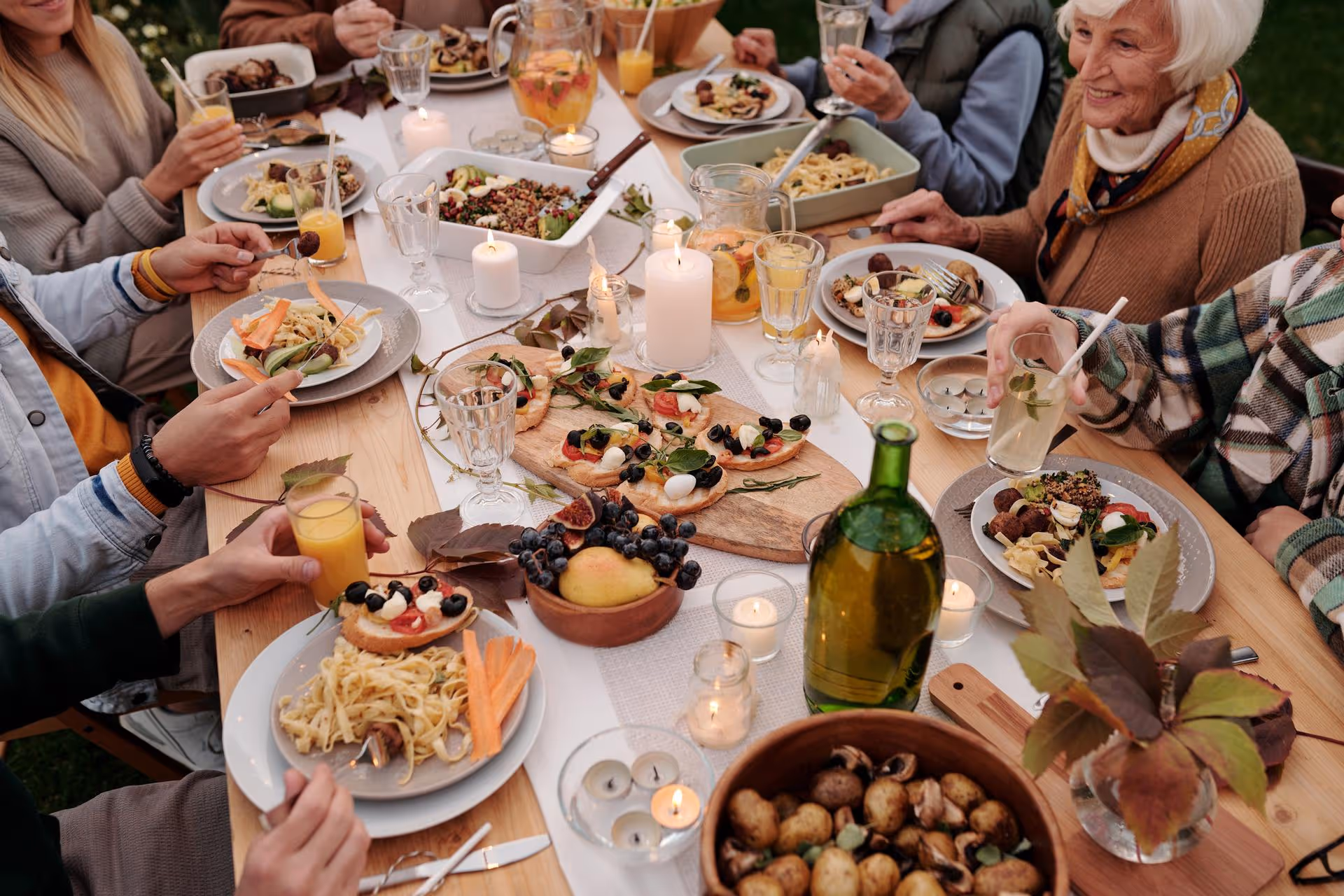 A diverse group of people enjoy an outdoor meal at a well-set wooden table, decorated with candles and autumn leaves, evoking a warm, festive atmosphere.