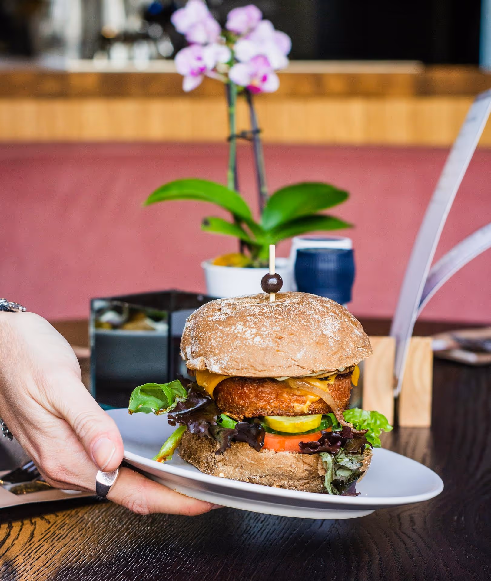 A hand places a veggie burger with lettuce, tomato, cheese, and a whole-grain bun on a table. In the blurred background, a pink orchid adds elegance.