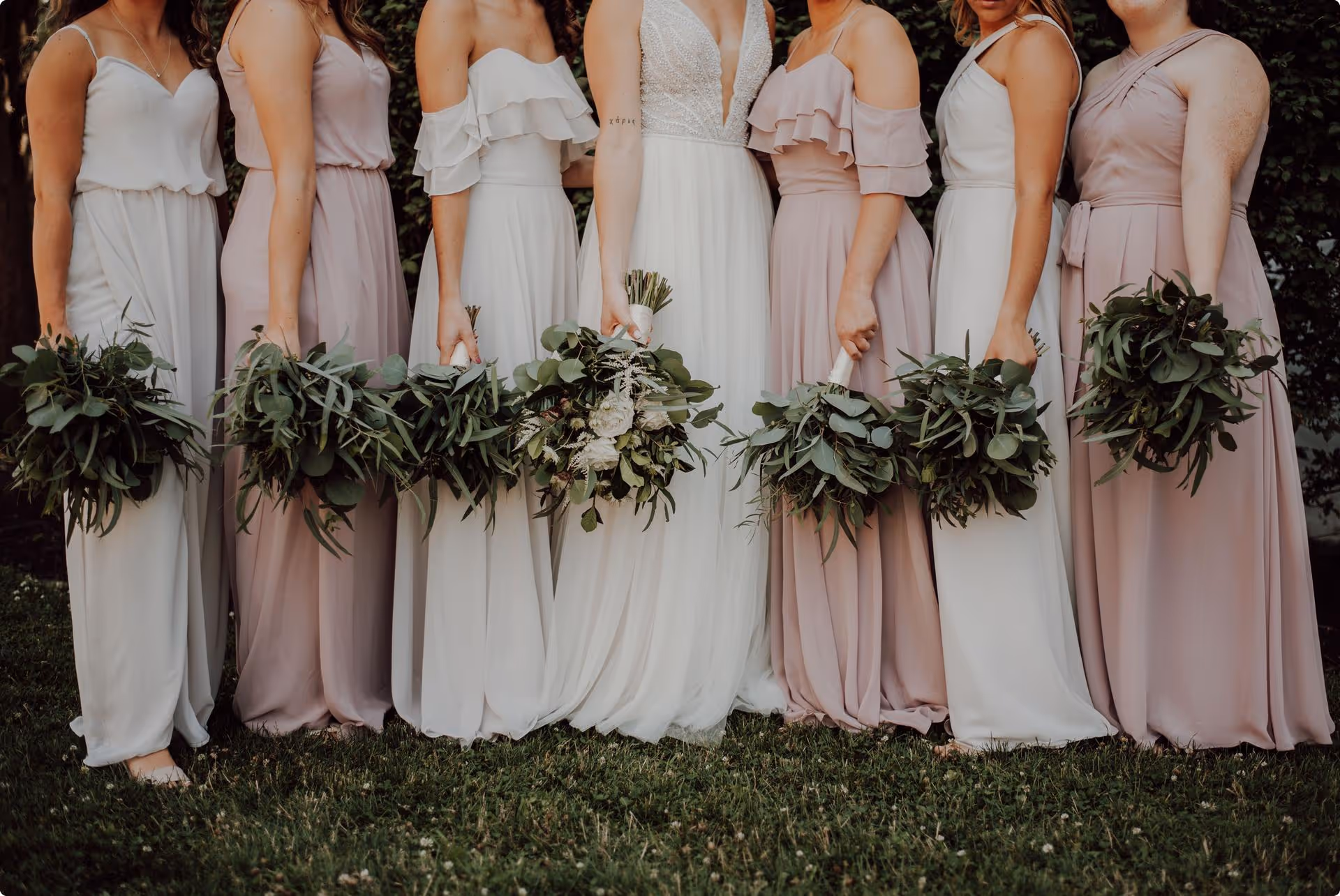 Bridesmaids in pastel dresses and holding leafy green bouquets stand beside the bride in a white gown, conveying a sense of elegance and celebration.
