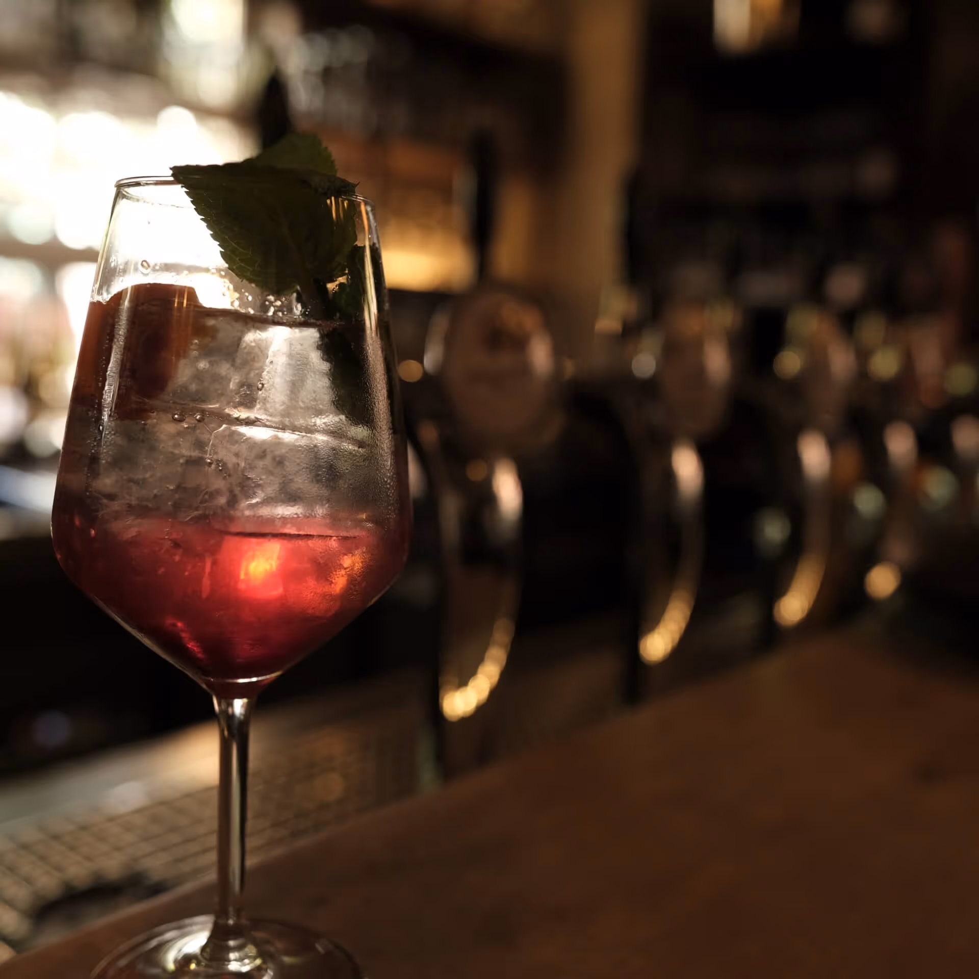 Glas mit rotem Getränk, Eiswürfeln und Minzblatt vor unscharfer Bar mit Zapfhähnen.