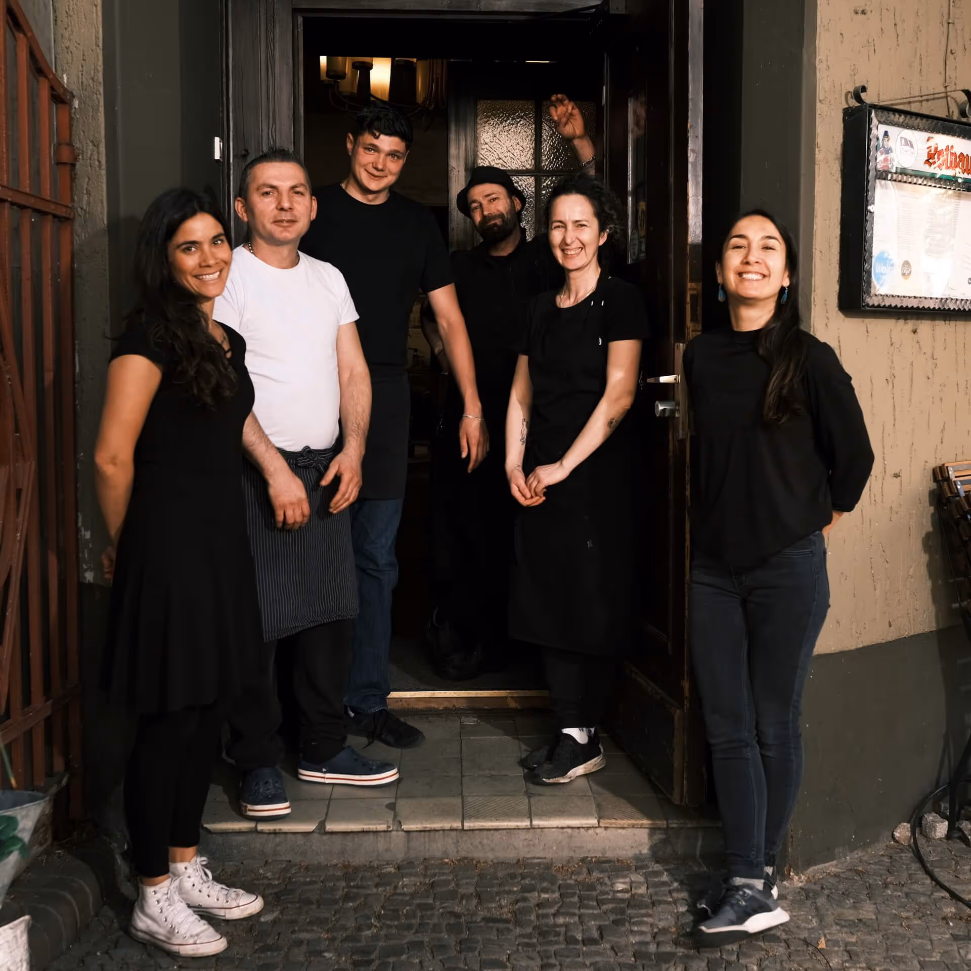 Sechs lächelnde Personen stehen vor offener Tür eines Gebäudes, Teamfoto draußen bei Tageslicht.