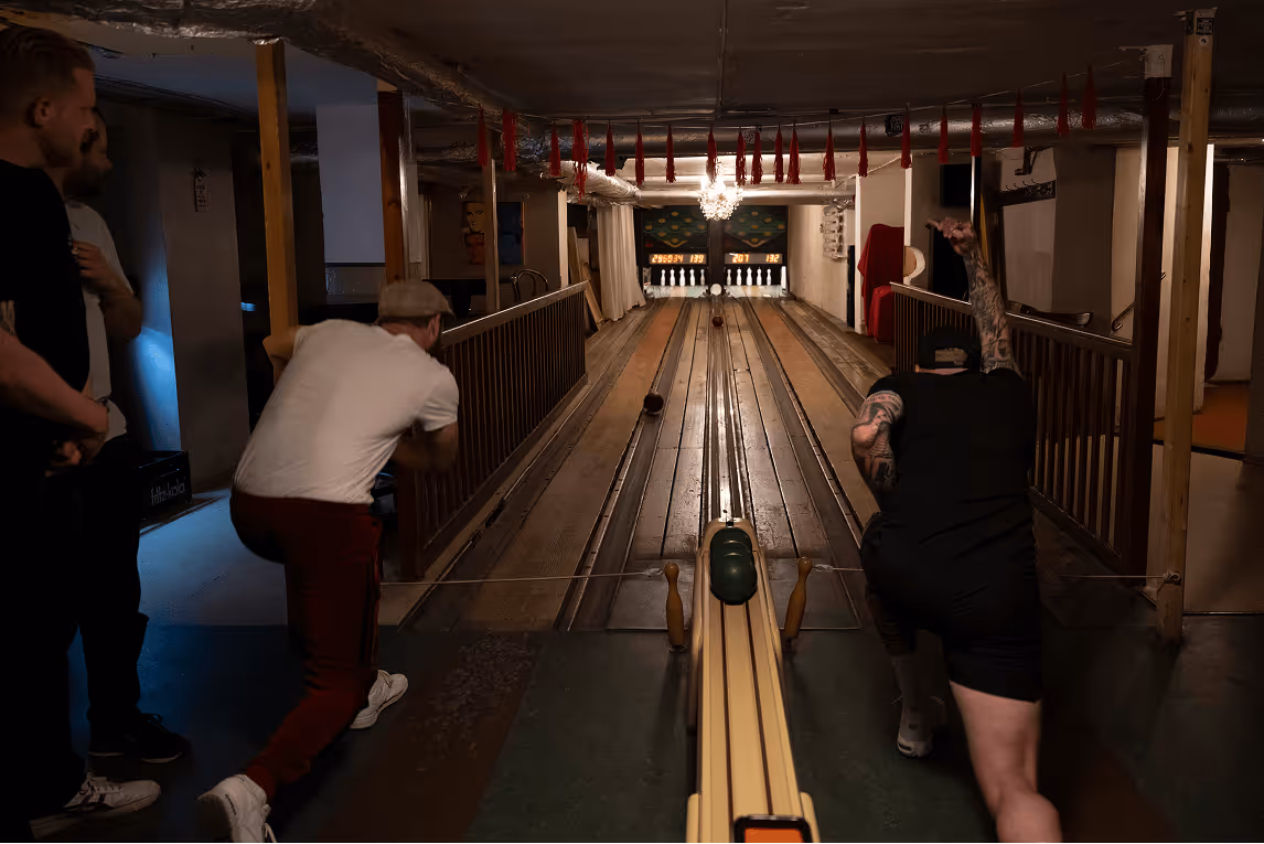 Zwei Männer werfen Bowlingkugeln auf einer Holzbahn, andere beobachten.