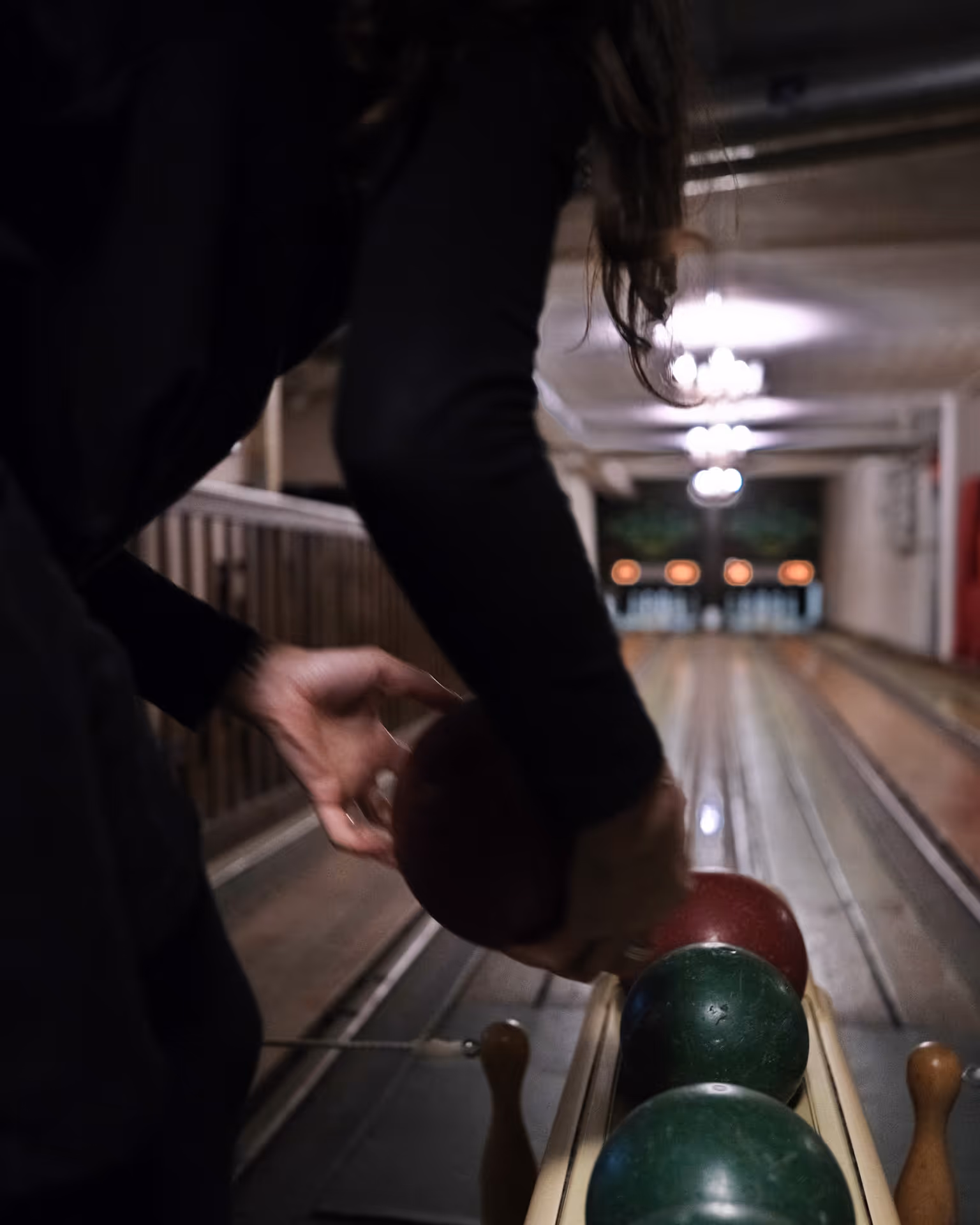 Person nimmt eine Bowlingkugel aus dem Regal vor einer Kegelbahn mit Holzdielen und Kegeln am Ende.
