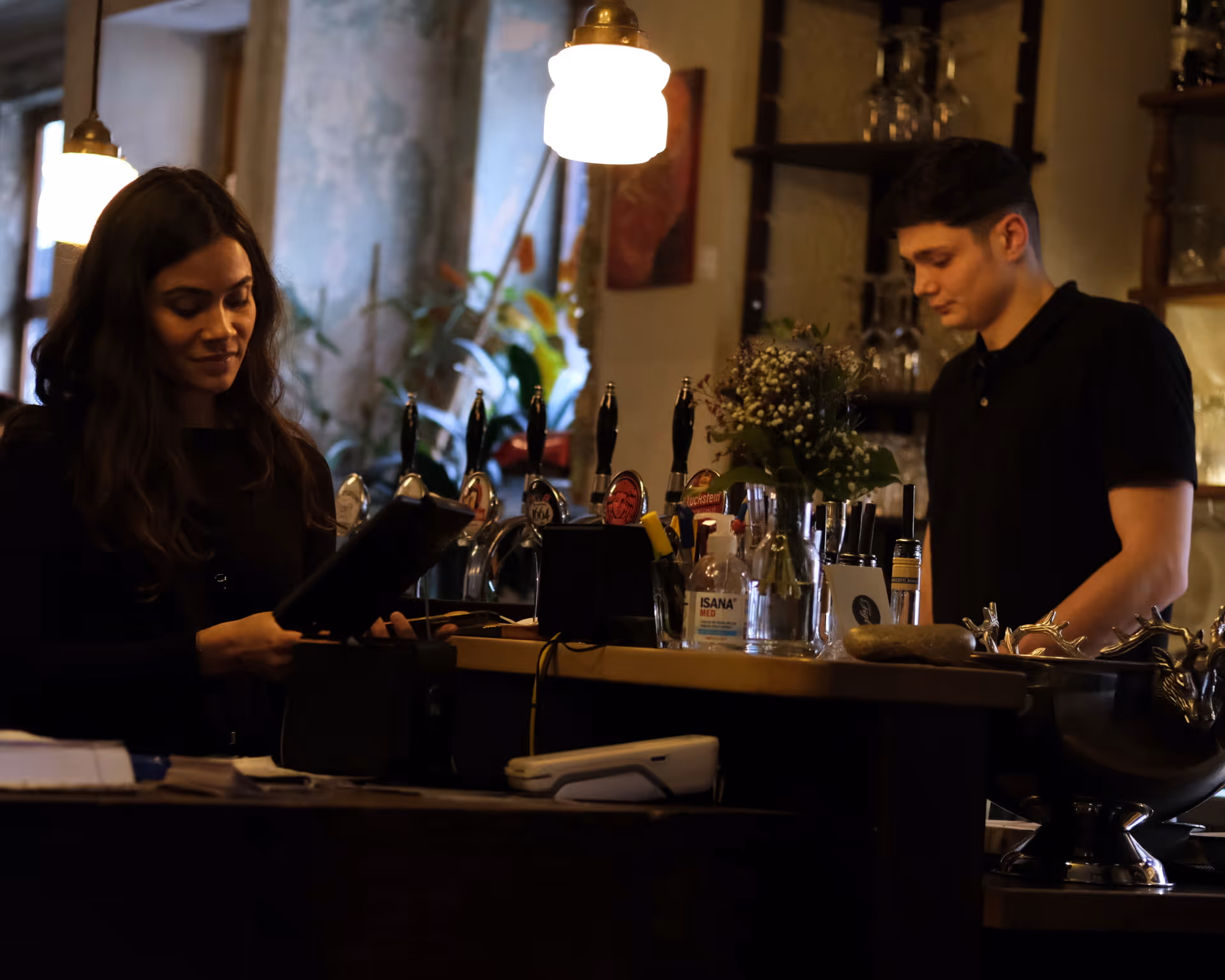 Eine Frau und ein Mann stehen an einer Bar mit Zapfhähnen, Blumen und einem Flaschenspender.