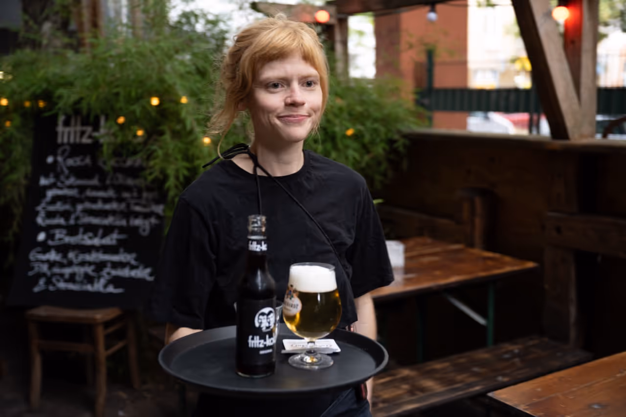 Junge Frau trägt ein Tablett mit einer Bierflasche und einem gefüllten Bierglas iim Außenbereich.